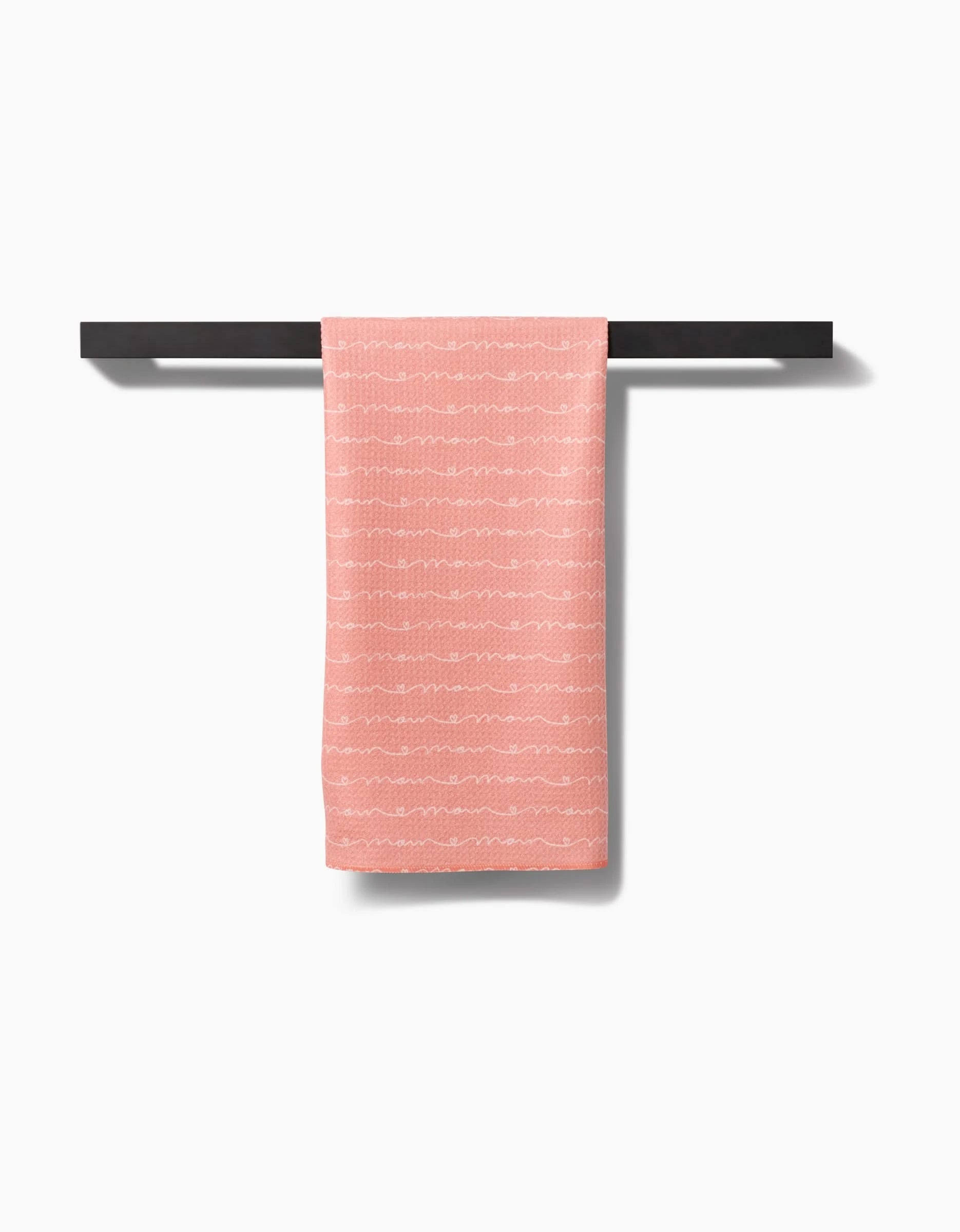 The “Love of Mom” tea towel by Sarah Reed, in light pink with subtle white wavy lines, is crafted from soft Geoweave™ fabric and hangs folded over a black towel bar against a white background.