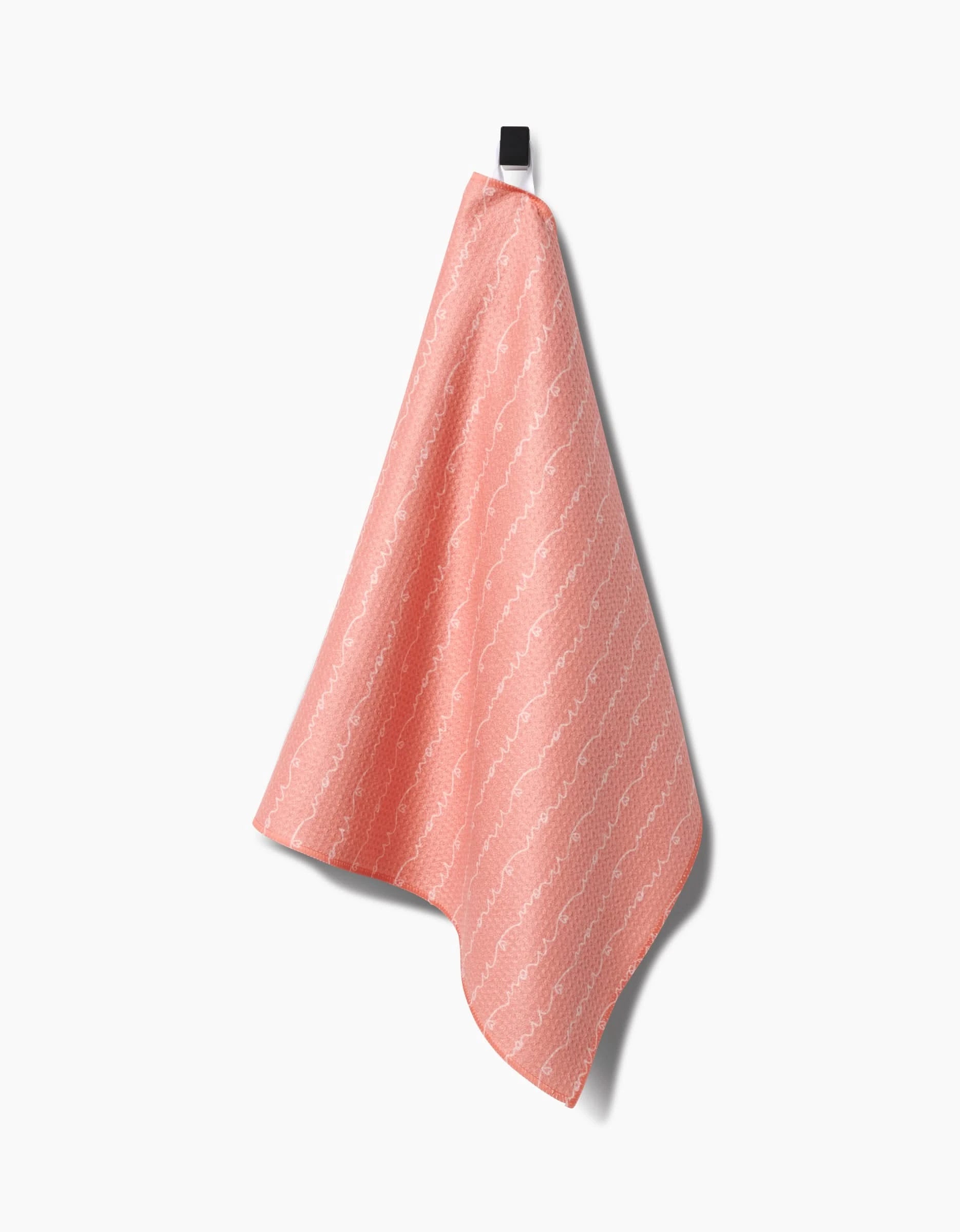 The Love of Mom Tea Towel by Sarah Reed, made from Geoweave™ fabric in coral pink with a white wavy pattern, hangs on a black hook against a plain white background.