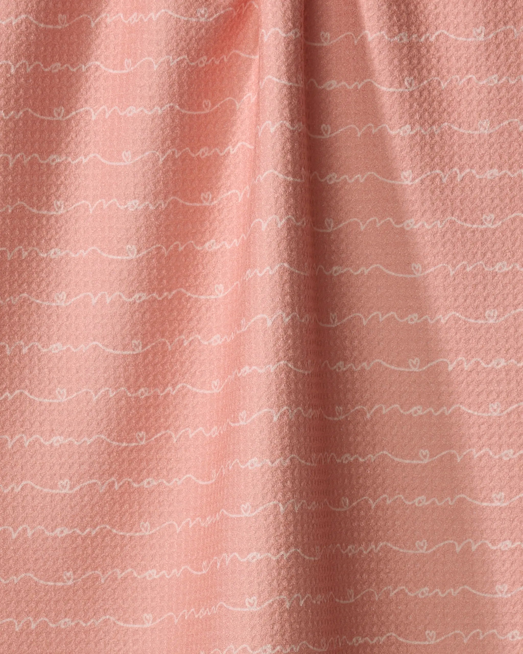 A close-up of the Love of Mom kitchen towel by Sarah Reed, featuring a soft pink, recycled fabric with white wavy lines and repeating cursive text on its gently textured, slightly gathered surface.