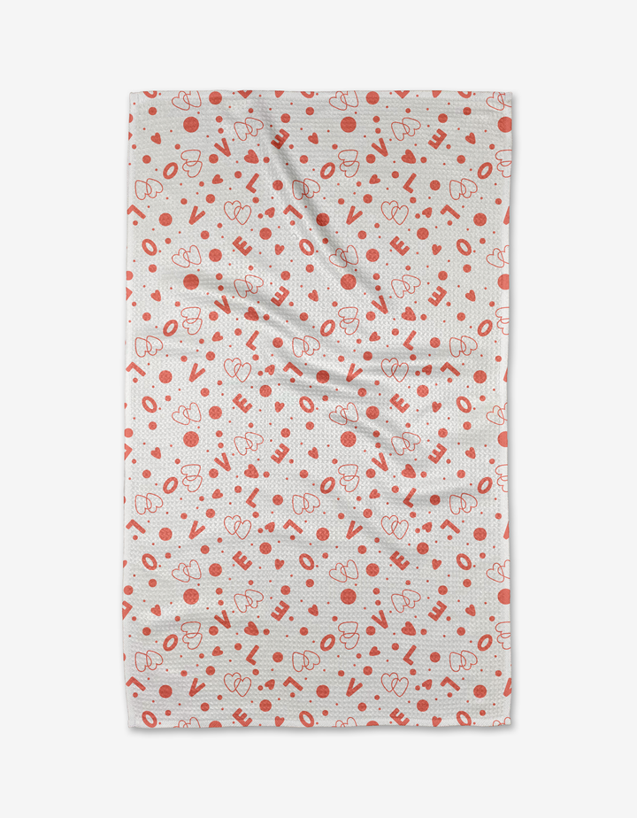 The Love Party kitchen towel by Charlie Rowan Designs is white, made from recycled materials, and features red hearts, dots, and abstract shapes. It appears slightly wrinkled and is shown against a plain white background.