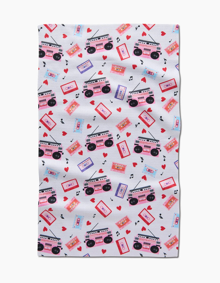 The Love Playlist tea towel by Vaishali Design Studio showcases pink and purple cassette tapes, boomboxes, red hearts, and black musical notes in a lively scattered pattern on white.