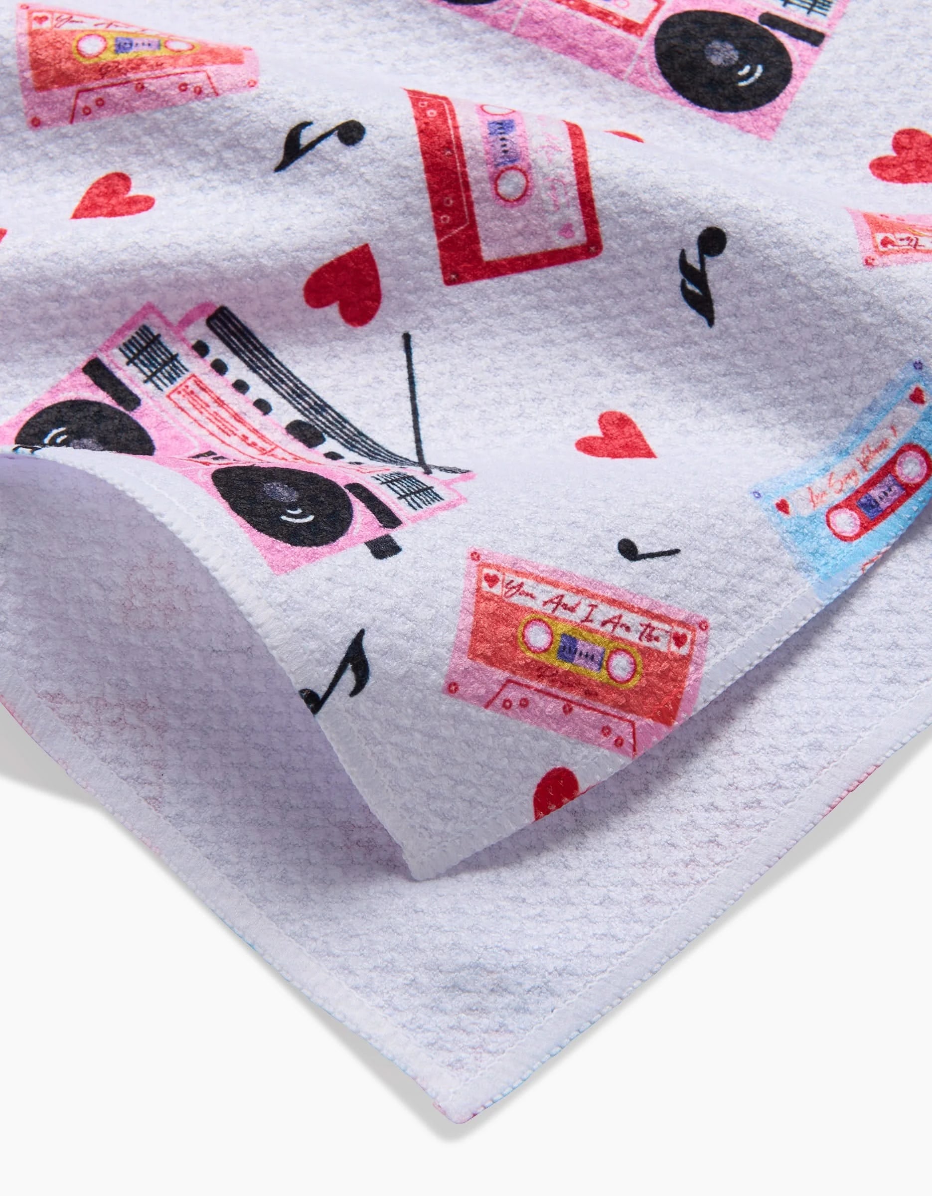 A close-up of the Love Playlist tea towel by Vaishali Design Studio, showcasing a textured white fabric with colorful cassette tapes, boomboxes, red hearts, and musical notes.