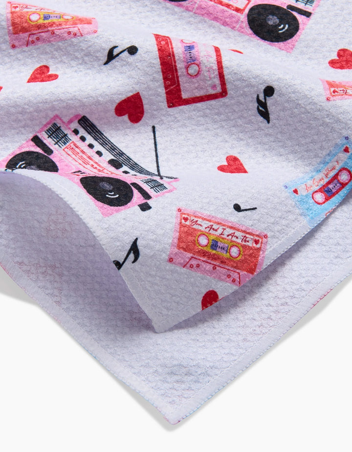 A close-up of the Love Playlist tea towel by Vaishali Design Studio, showcasing a textured white fabric with colorful cassette tapes, boomboxes, red hearts, and musical notes.