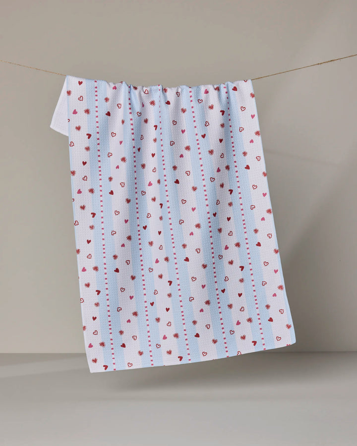 Geometry's Love Stripes tea towel, made from recycled materials, features small red and black hearts with blue dotted stripes and is shown hanging against a light background.