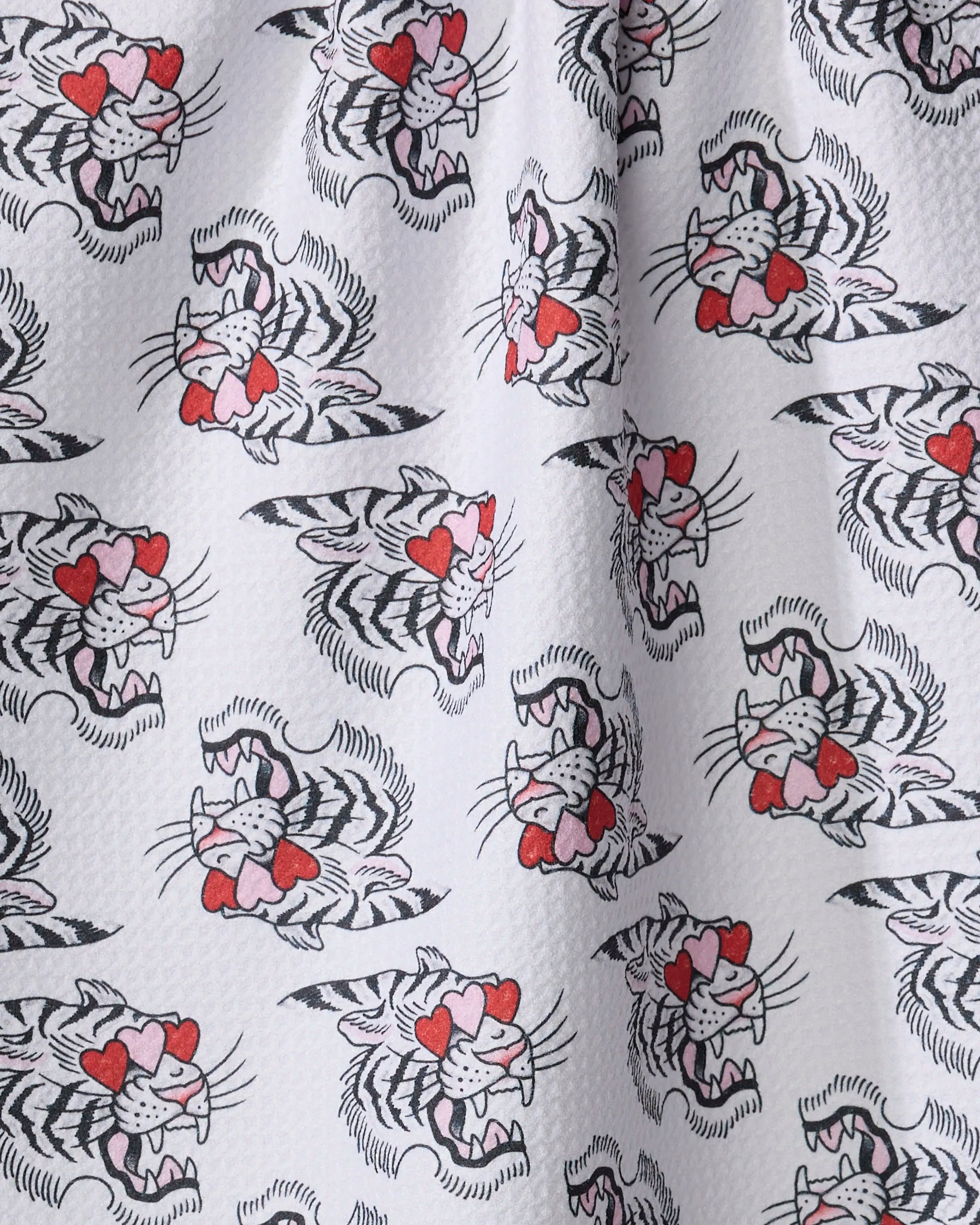 The Cole Strem Love Tiger tea towel features white fabric with a repeating cartoon tiger pattern—heart-shaped eyes, red noses, and snarling faces. Ultra-absorbent and made from recycled materials, it has a slightly dimpled texture.