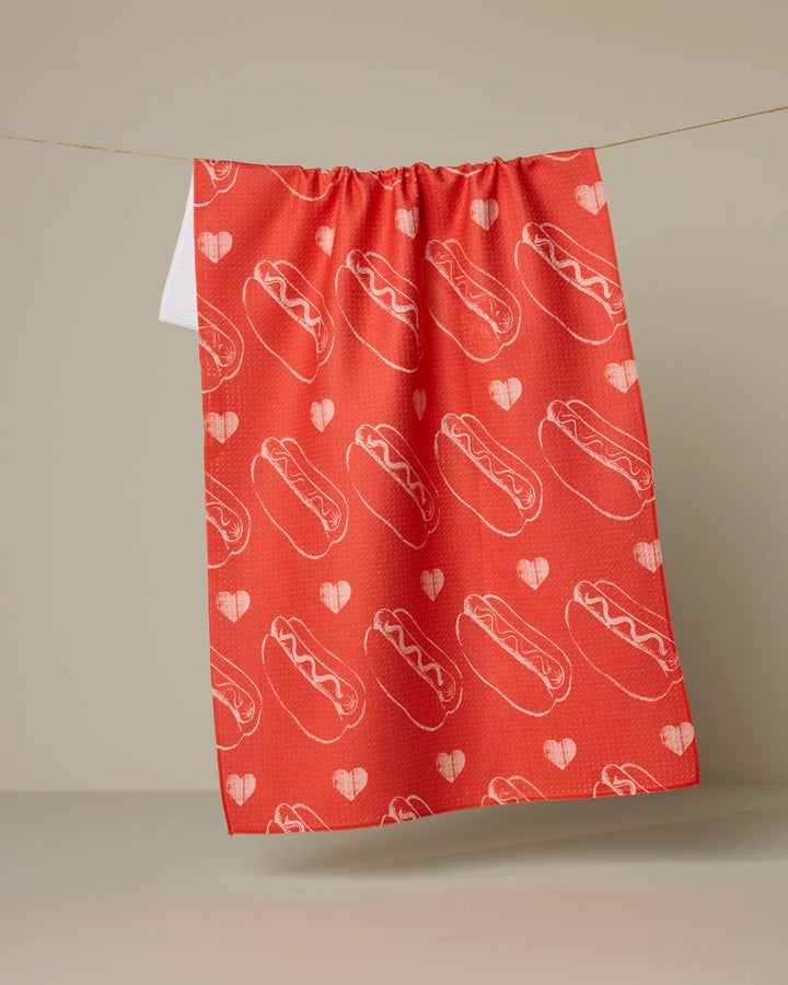 The Geometry "Lovely Franks" kitchen towel, made from recycled materials in red, features a repeating pattern of outlined hot dogs and hearts and hangs on a string against a neutral background.