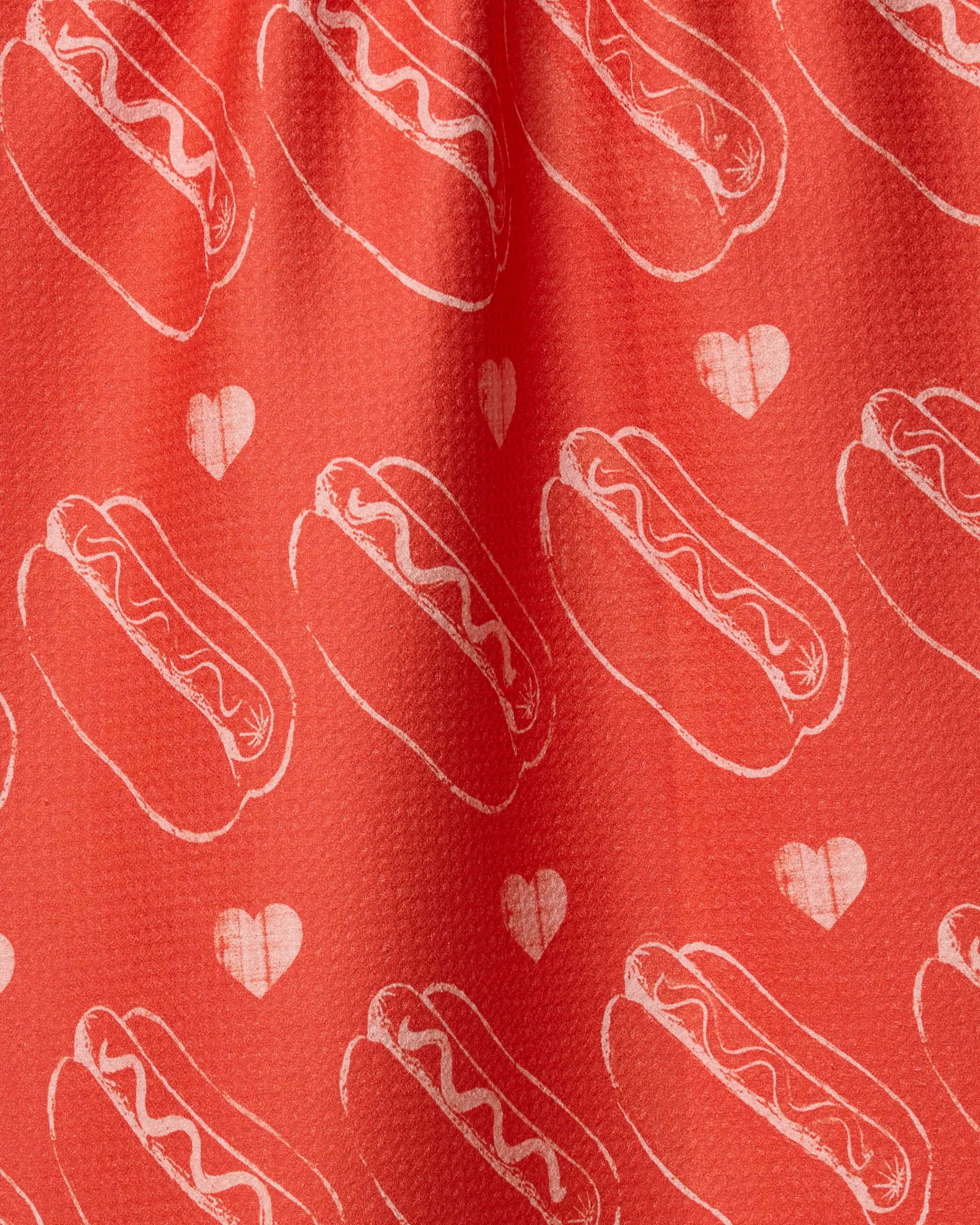The Geometry Lovely Franks recycled kitchen towel features red fabric with white hot dog and heart outlines in an even, repeating pattern. Slight gathers create soft folds and subtle shadows across its surface.