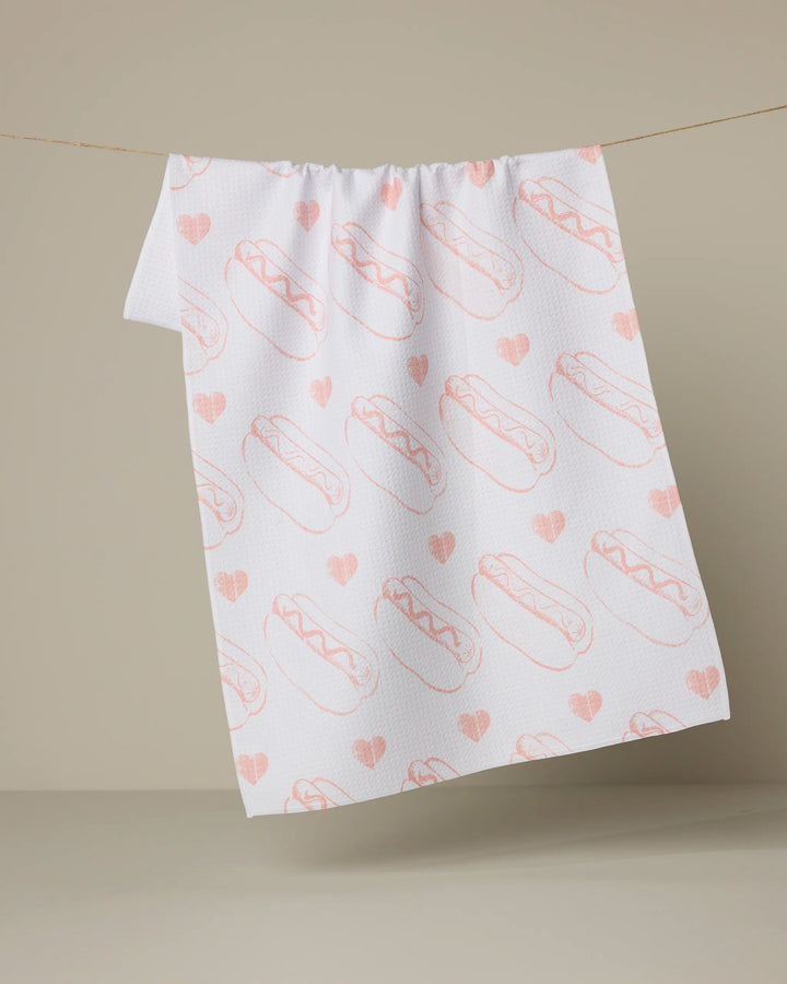 A white recycled kitchen towel by Geometry, called Lovely Franks, hangs on a line against a neutral background and features light pink hot dogs and hearts.