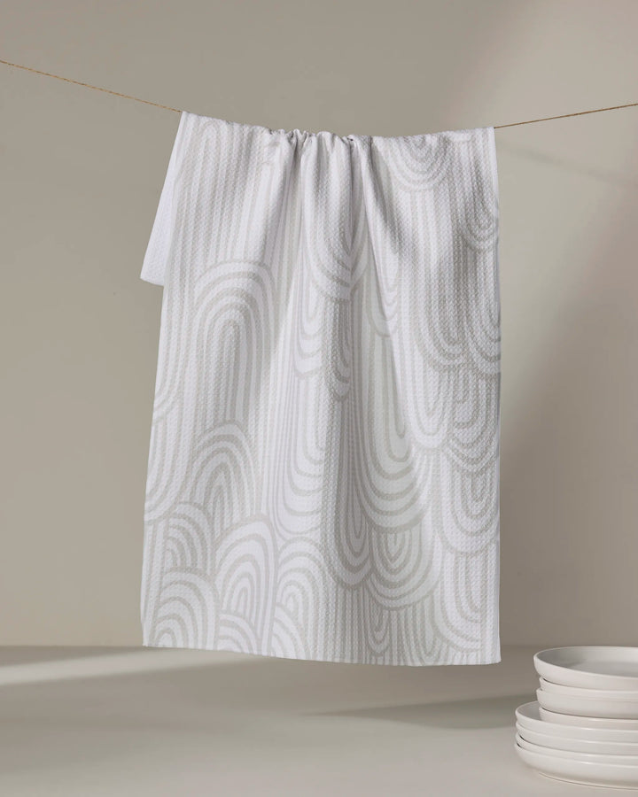 A recycled kitchen towel, Lovers Arc by Hufton Studio, in white with a light gray abstract wave pattern hangs on a line. A small stack of white bowls sits on a neutral surface in the lower right corner.