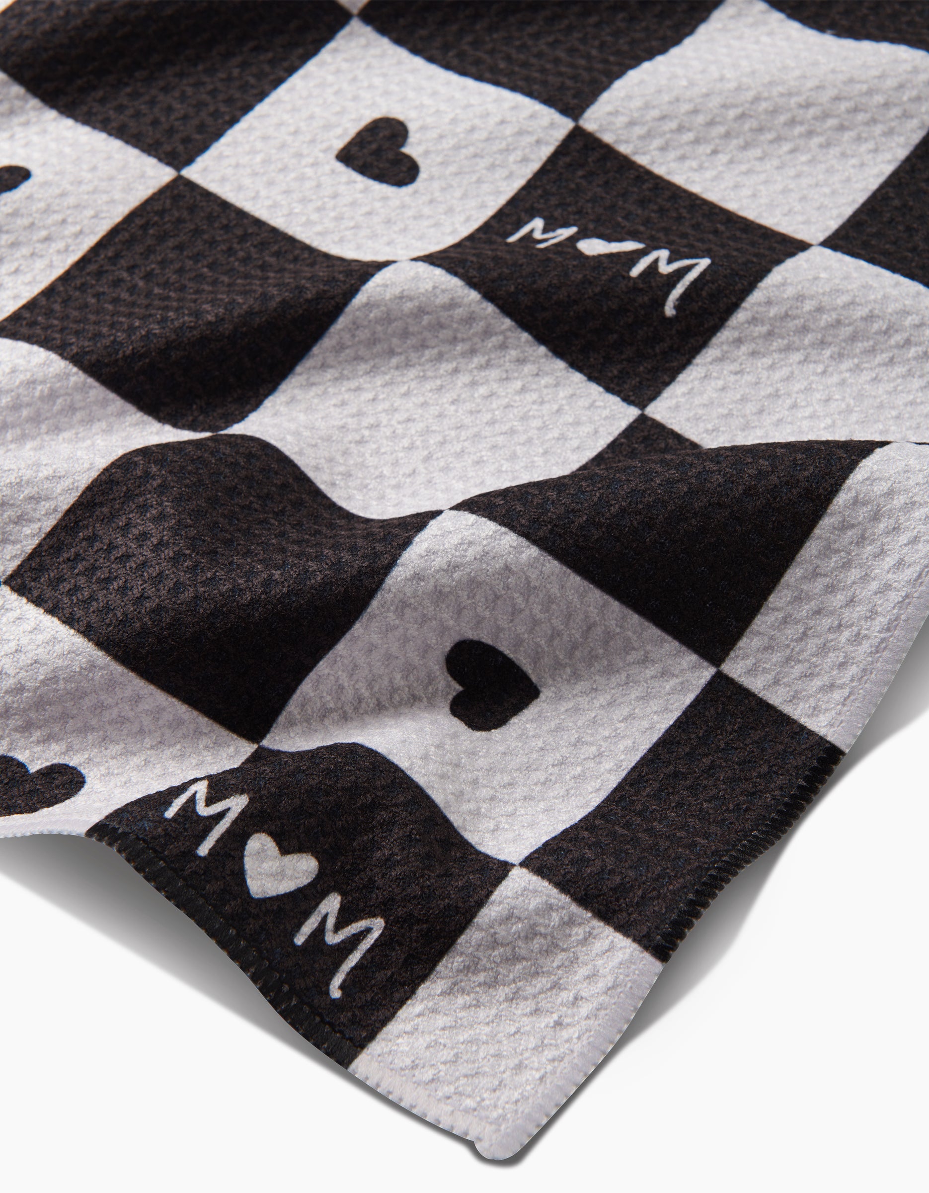 A close-up of GEOMETRYs Loving Tea Towel in black and white checkered Geoweave fabric, featuring heart shapes and MOM text in some squares. The textured surface appears soft, and the towel is slightly folded.