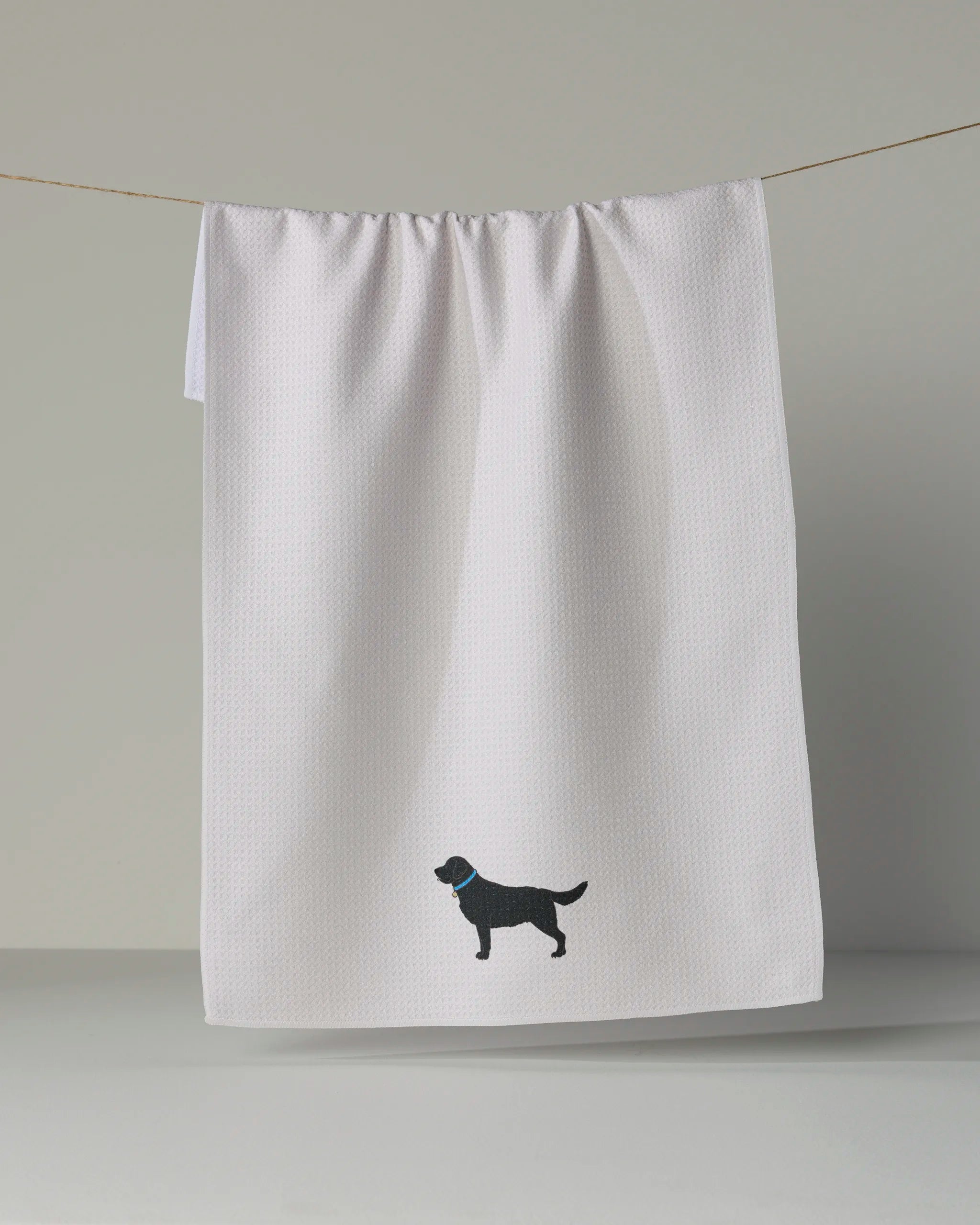 A white tea towel from Geometry called “Loyal Lab” hangs on a line, featuring a small black labrador silhouette with a blue collar near the bottom center.