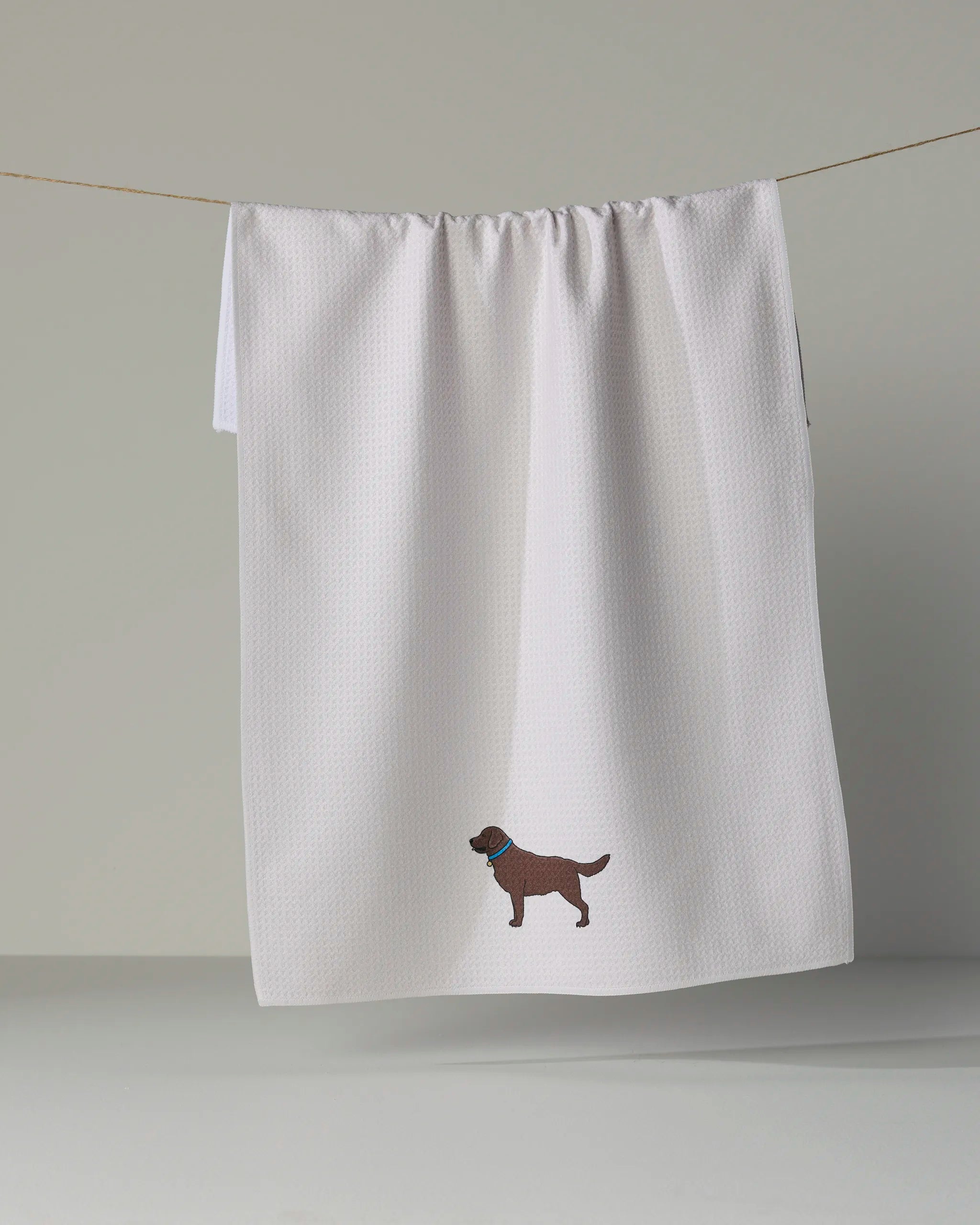 A white “Loyal Lab” kitchen tea towel by Geometry hangs on a line, featuring an embroidered brown dog wearing a blue collar near the bottom edge.