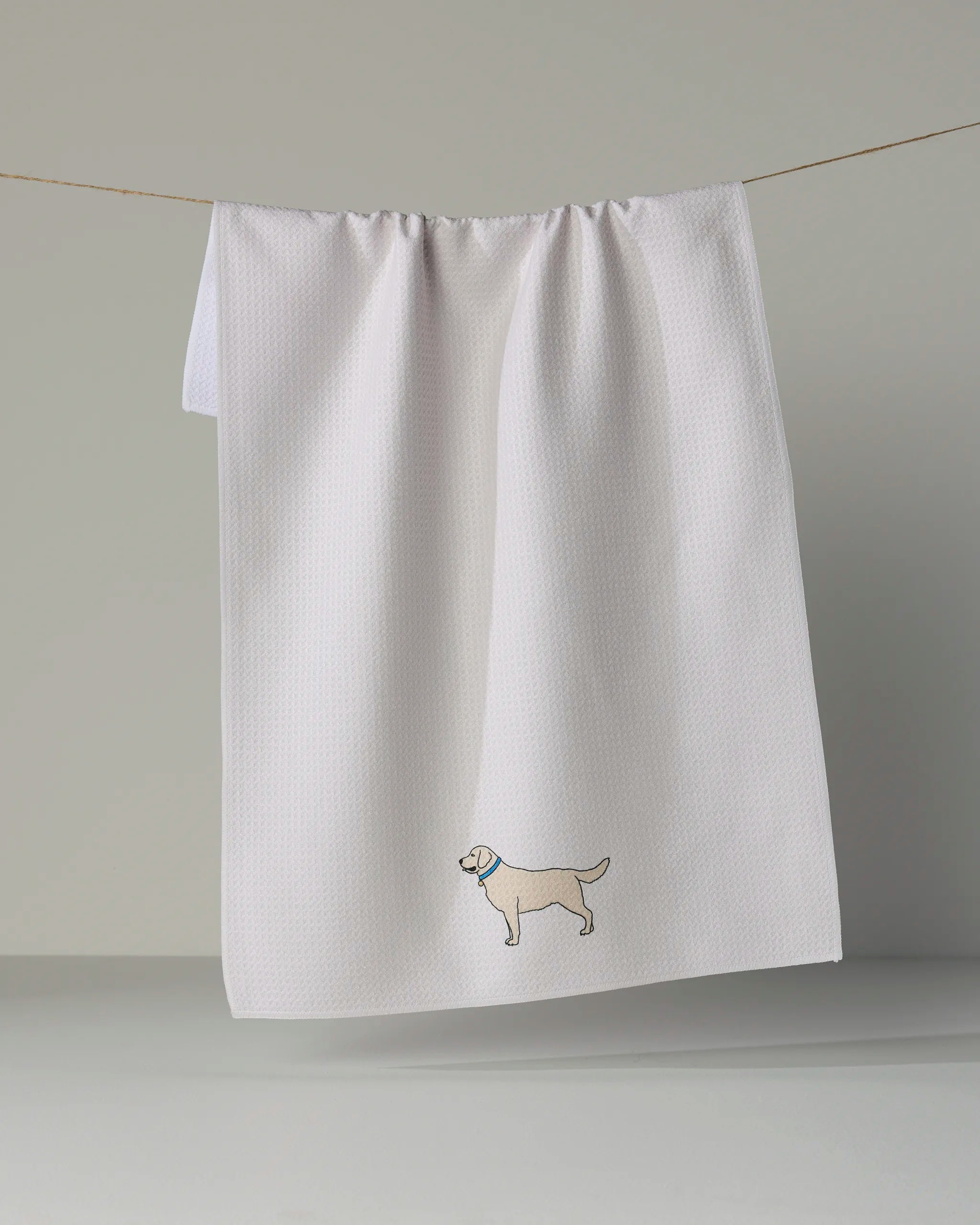 A white textured “Loyal Lab” kitchen tea towel by Geometry hangs on a line, featuring a small, embroidered yellow dog near the bottom corner.