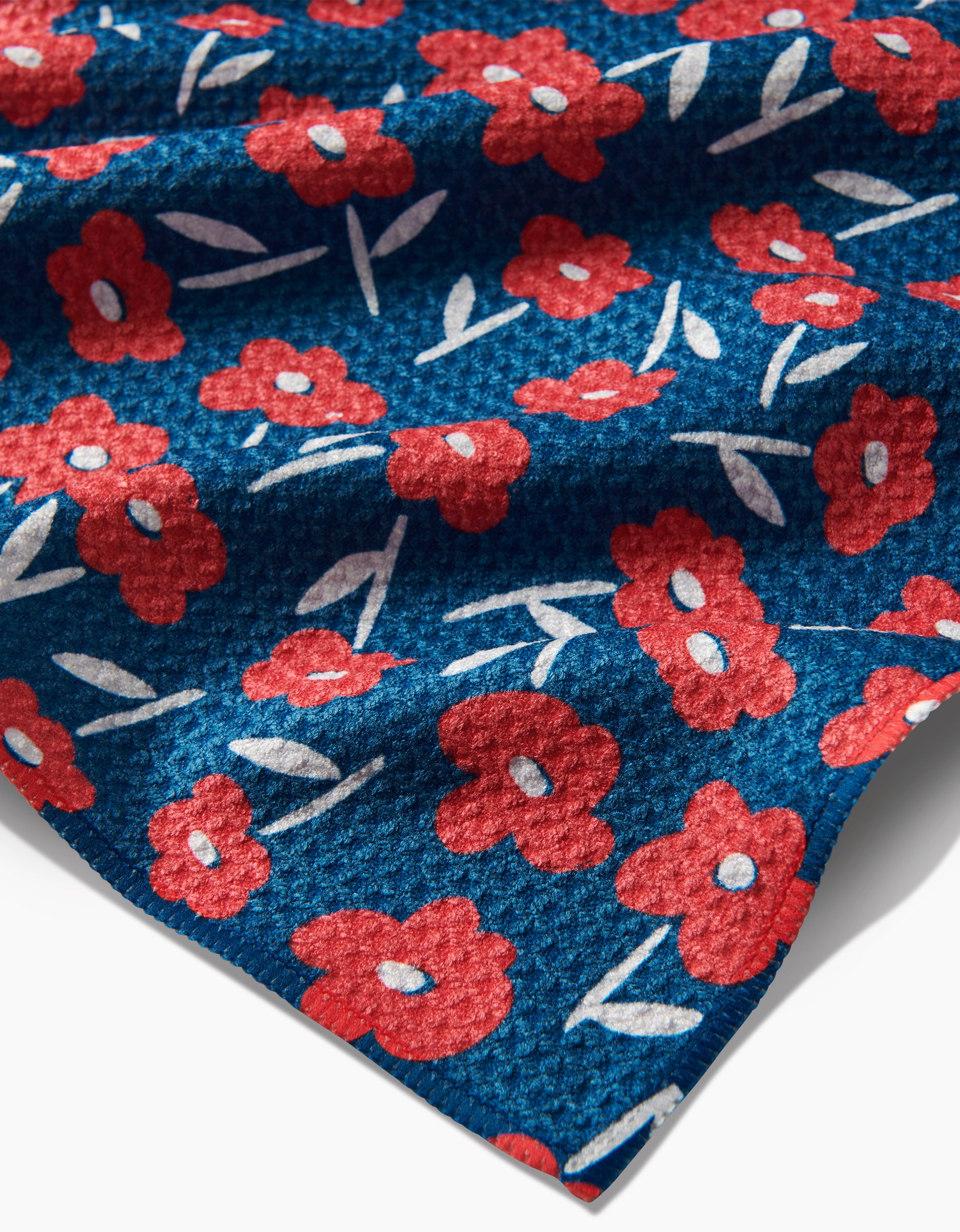 A close-up of Hufton Studio’s Luca Daisy Red tea towel shows blue Geoweave fabric with a red daisy and white leaf pattern. Made from recycled materials, the cloth features soft folds and shadows from gentle wrinkling.