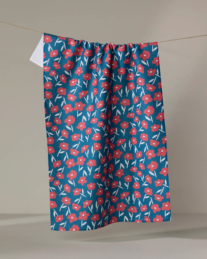 The Hufton Studio Luca Daisy Red tea towel, featuring a red and white floral design on blue, is displayed hanging against a plain beige background.