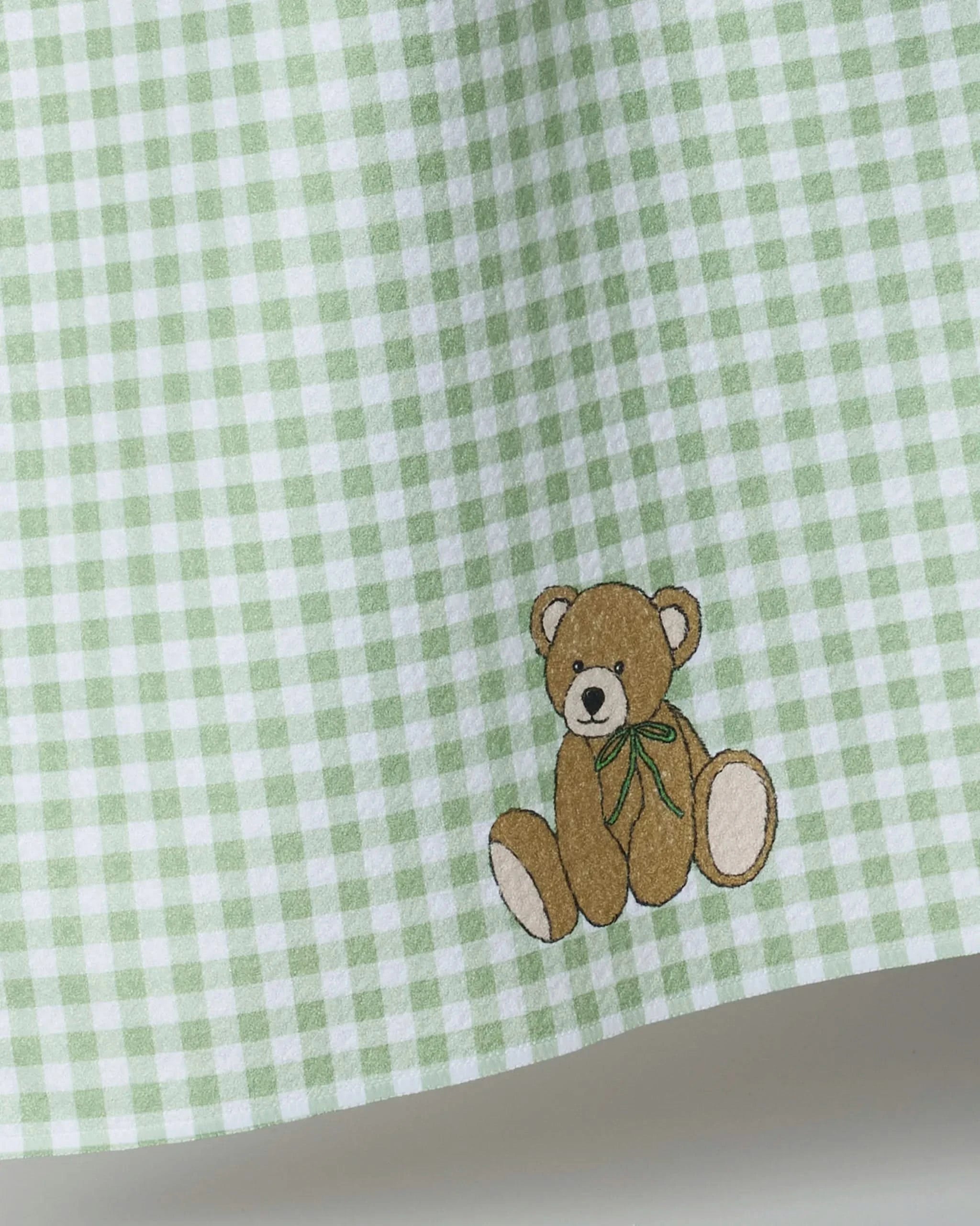 A close-up of Geometry’s Lucky Bear tea towel showcases green and white checkered fabric with an embroidered brown teddy bear with a green ribbon near the bottom right corner.
