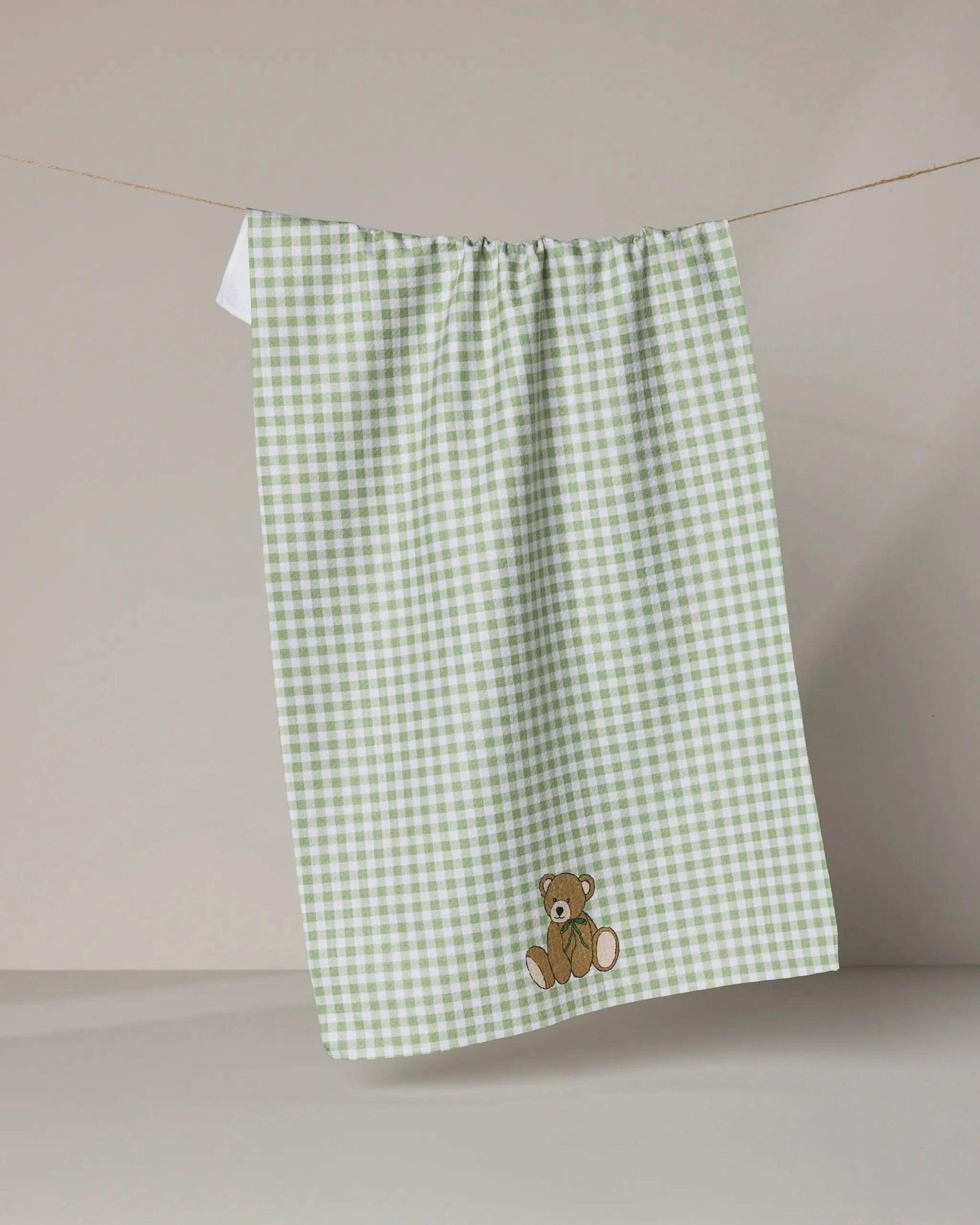 The Geometry Lucky Bear kitchen towel features green and white gingham with an embroidered brown teddy bear, shown hanging on a clothesline against a beige background.
