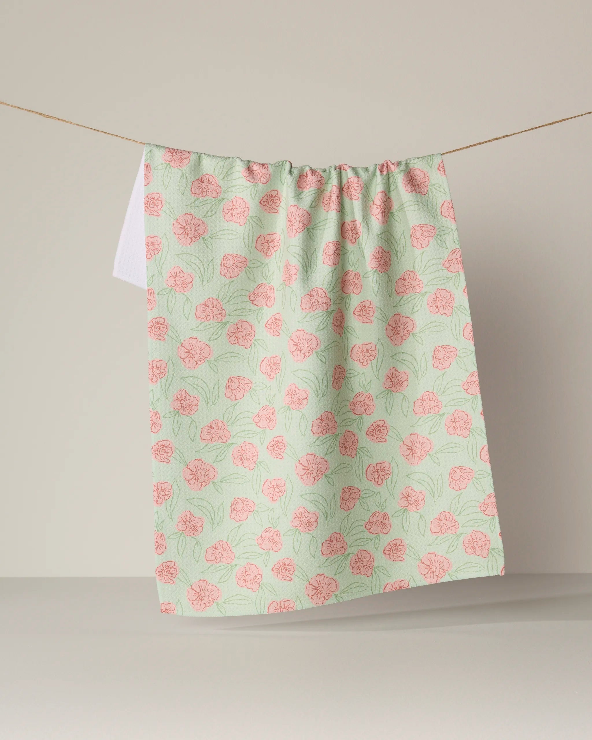 A pastel green Geometry "Lucky Bloom" kitchen tea towel, featuring a pink floral pattern and made from recycled materials, hangs on a thin string against a neutral background.