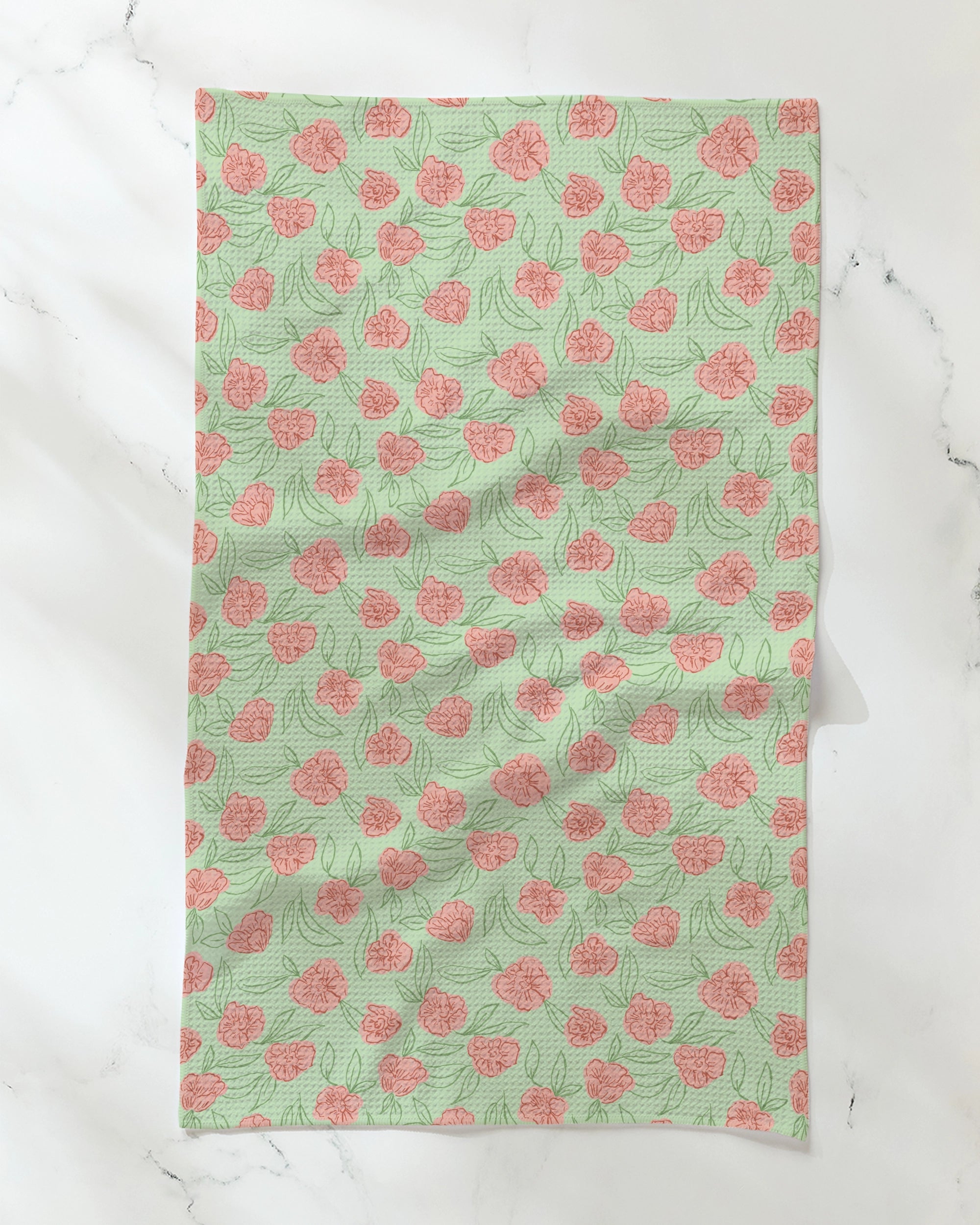 A rectangular tea towel from Geometry, called Lucky Bloom, featuring a light green background with a repeating pink floral pattern, is laid flat on a white marble surface. Made from recycled materials, the fabric displays subtle wrinkles.