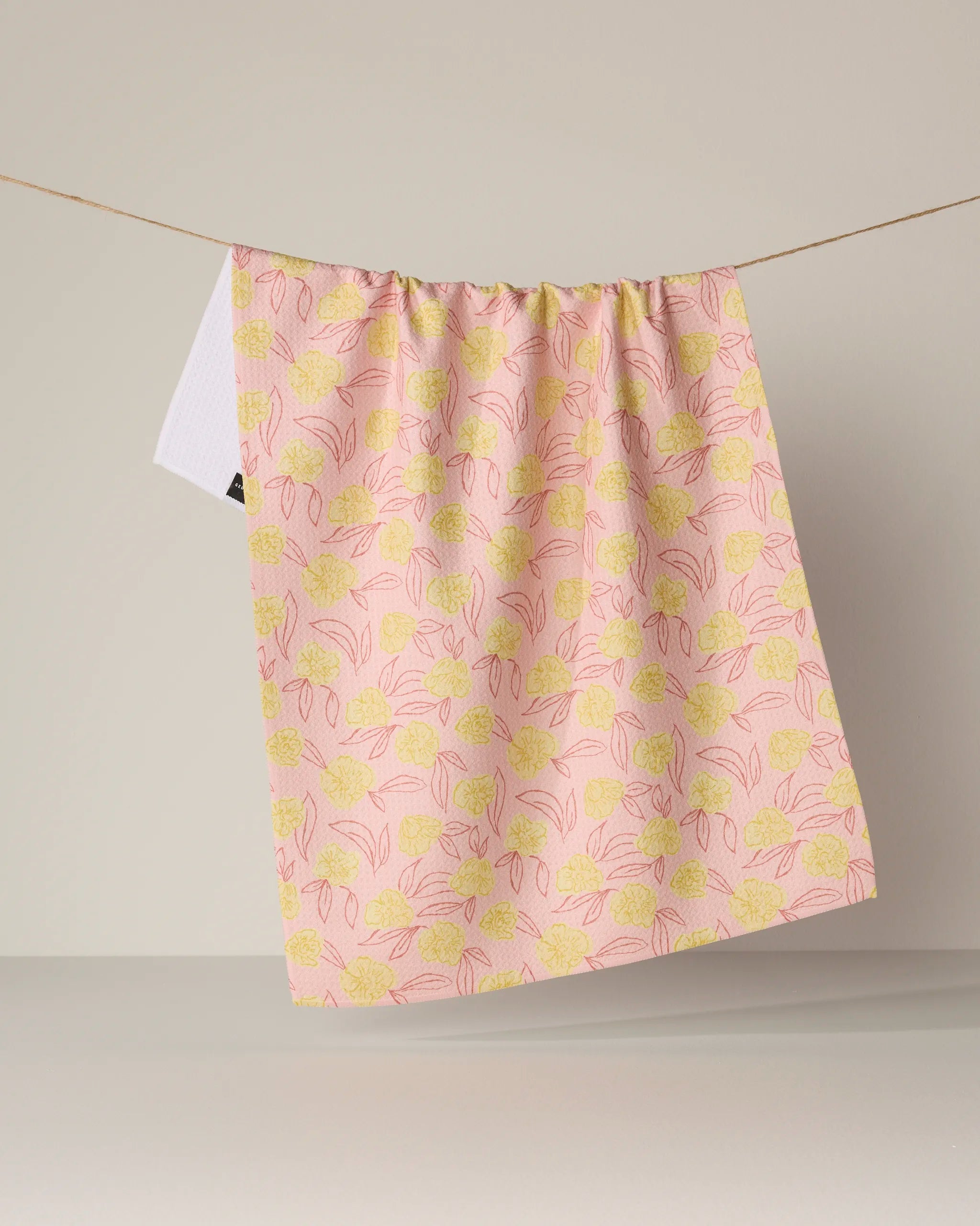 The Geometry Lucky Bloom kitchen tea towel, featuring a pink and yellow floral pattern, hangs on a string against a plain beige background and is crafted from recycled materials for an eco-friendly touch.