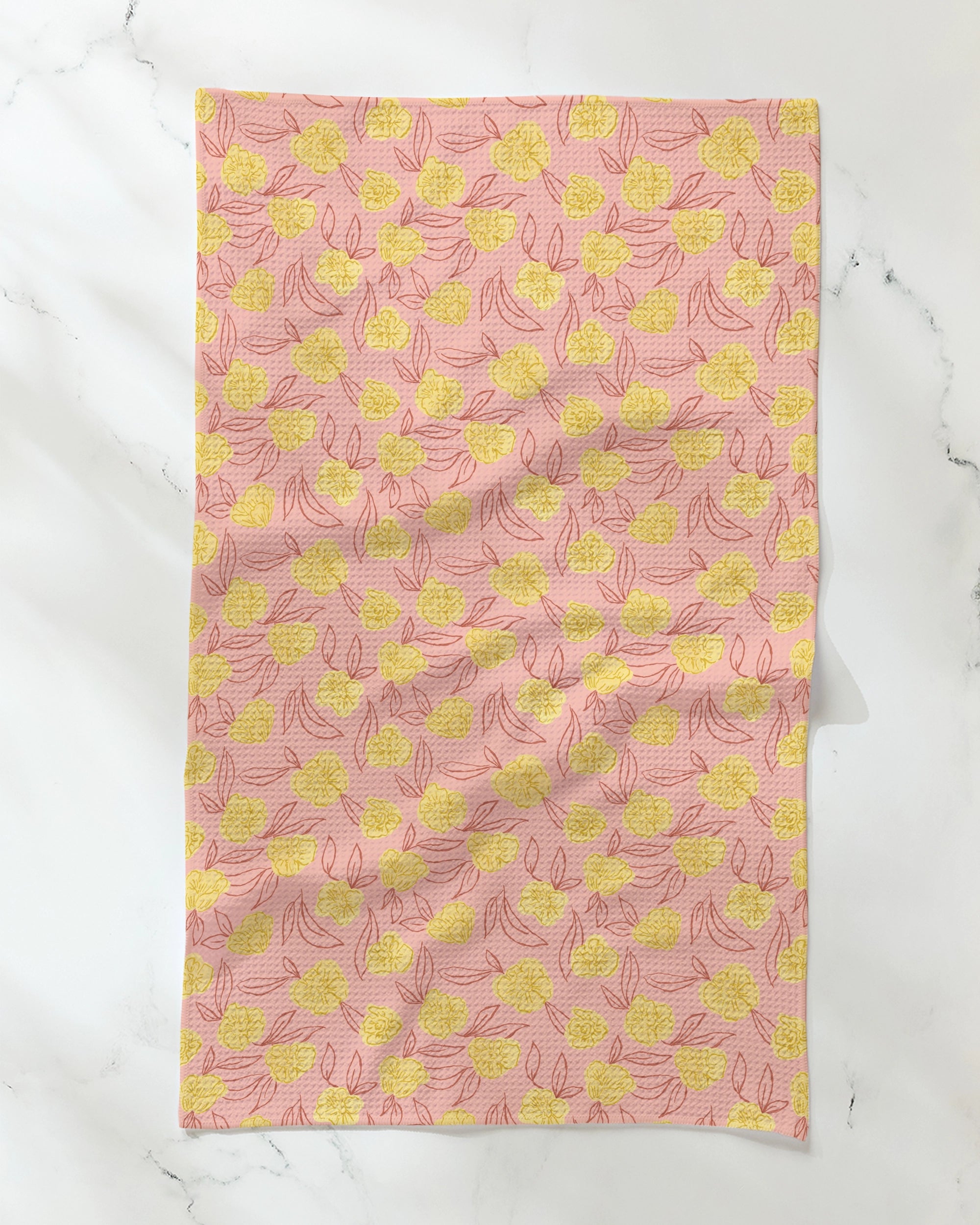 A Geometry Lucky Bloom tea towel, featuring a yellow floral pattern on a pink background, is laid flat on a white marble surface. Made from ultra-absorbent recycled materials, the rectangular fabric appears slightly wrinkled.