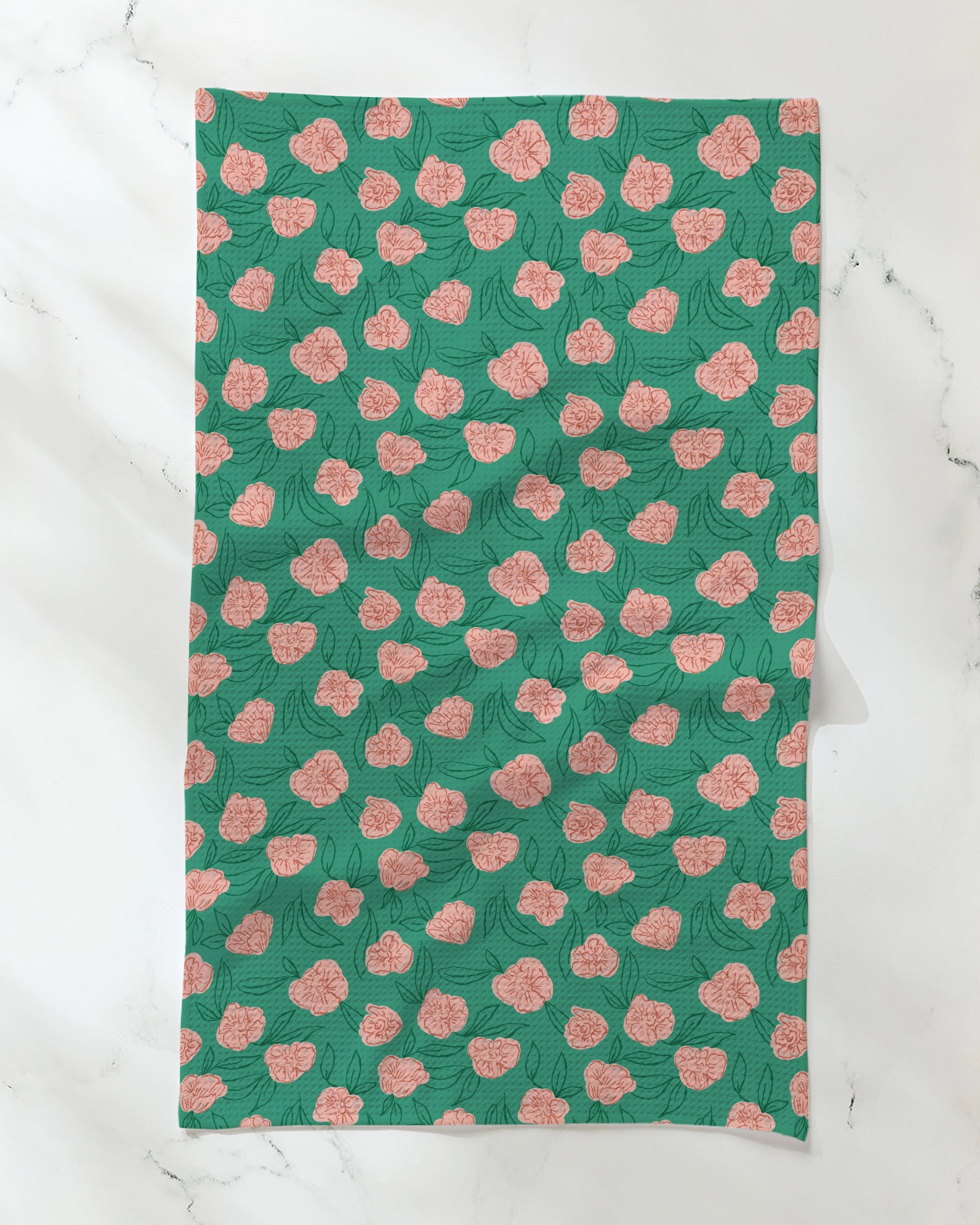 The Lucky Bloom tea towel by Geometry, featuring a repeating pattern of pink flowers and green leaves on ultra-absorbent recycled material, is displayed with visible creases on a white marble surface.