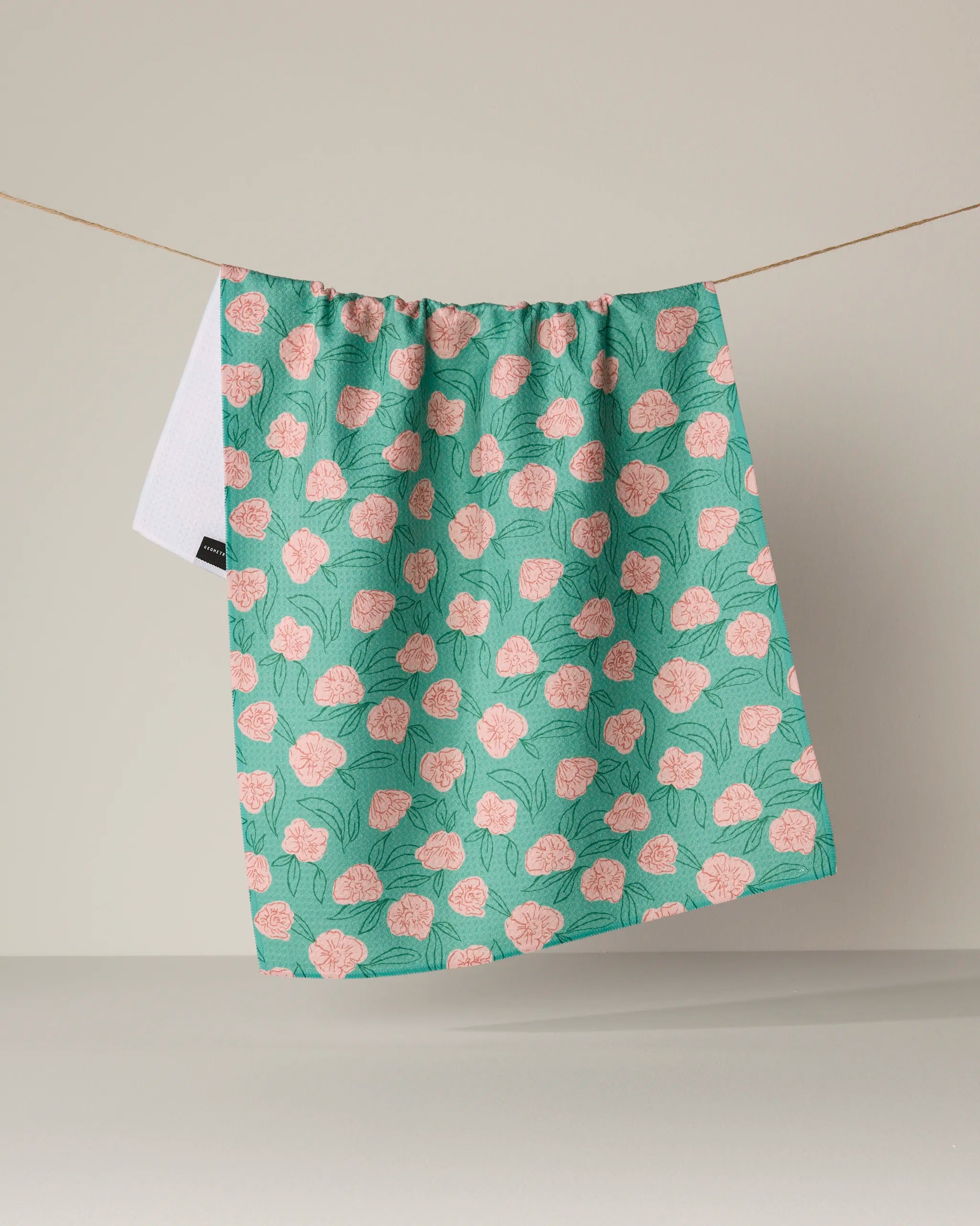 The "Lucky Bloom" kitchen tea towel by Geometry, featuring a green background with a pink floral pattern and made from recycled materials, hangs on a string against a neutral backdrop.
