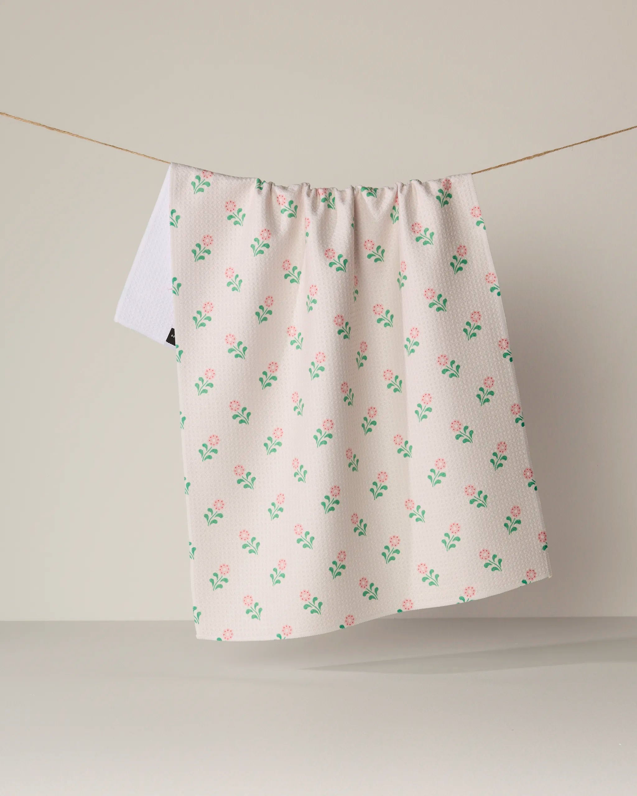 The Lucky Blossom kitchen towel by Geometry, featuring a pink and green floral pattern on white and crafted from recycled materials, hangs on a clothesline against a plain beige background.