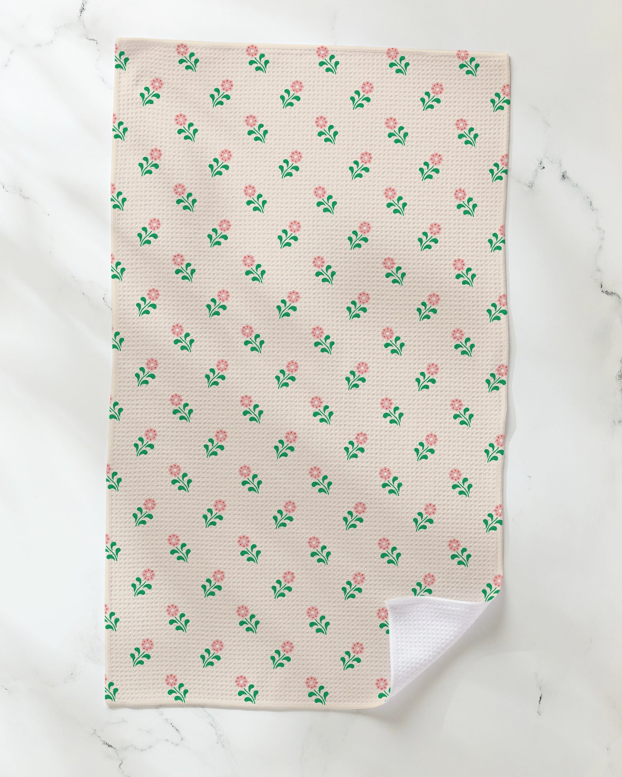 The Lucky Blossom quick-drying dish towel by Geometry features a beige background adorned with small pink flowers and green leaves. Displayed on a white marble surface, one corner of the towel is slightly turned up to reveal its white reverse side.