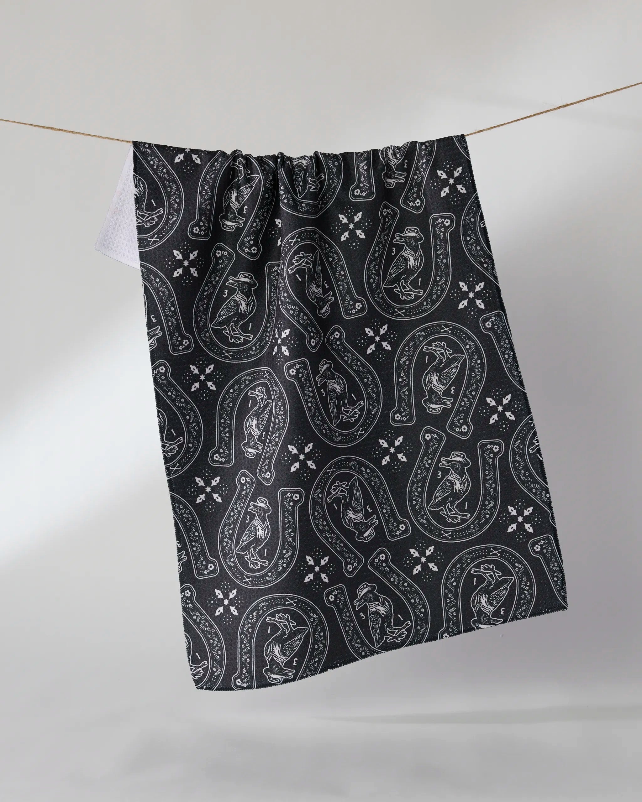 A Geometry "Lucky Crow" ultra-absorbent kitchen towel hangs on a clothesline, featuring black-and-white outlined horseshoes, horse heads, and floral details on a dark background.