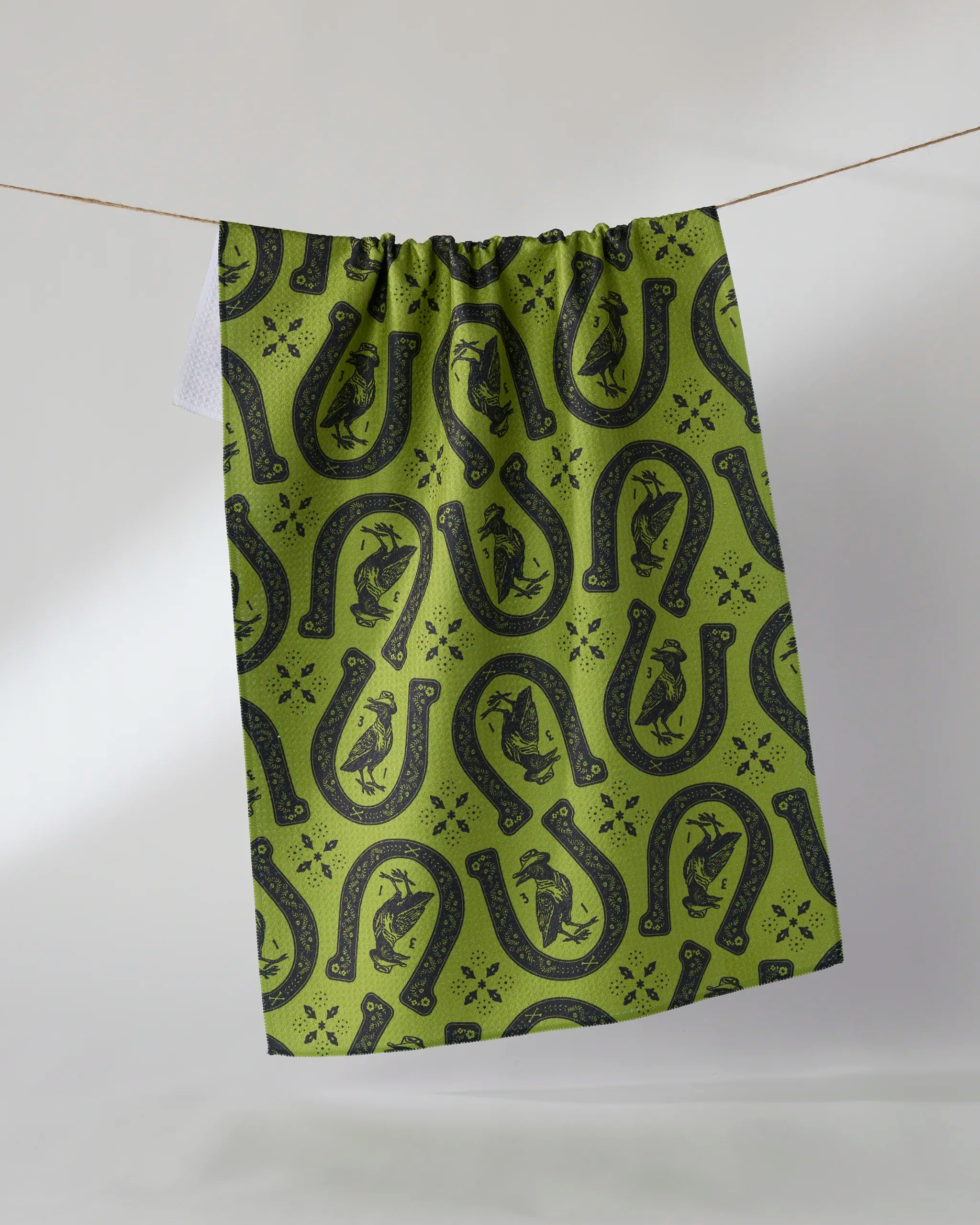 Geometry's Lucky Crow kitchen towel, crafted from recycled materials, features a green fabric with black horseshoes and horse patterns, hanging on a clothesline against a simple light background.