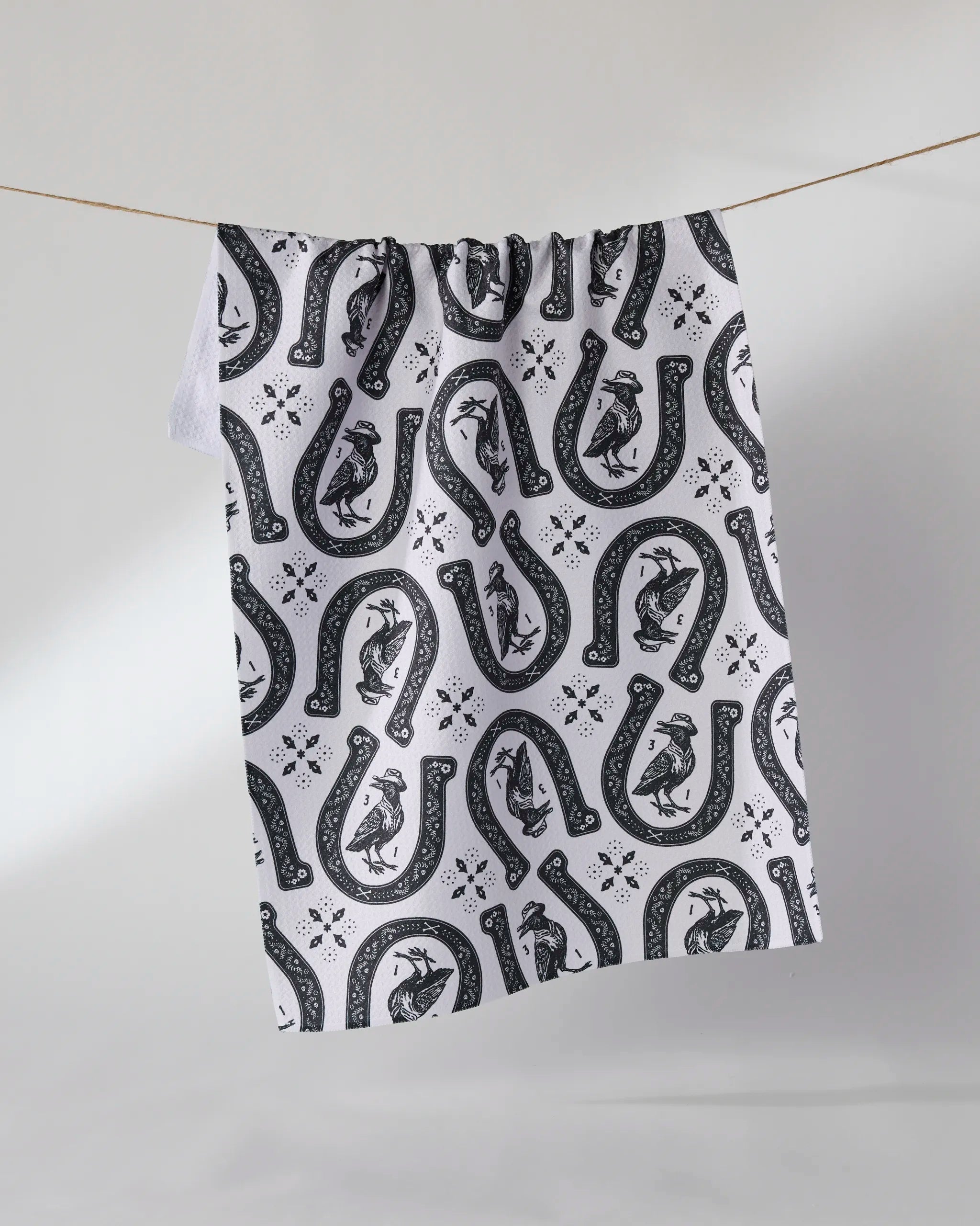 The Geometry Lucky Crow kitchen towel, featuring a black pattern of horseshoes, birds, and flowers on ultra-absorbent white fabric, hangs on a clothesline against a plain light background.