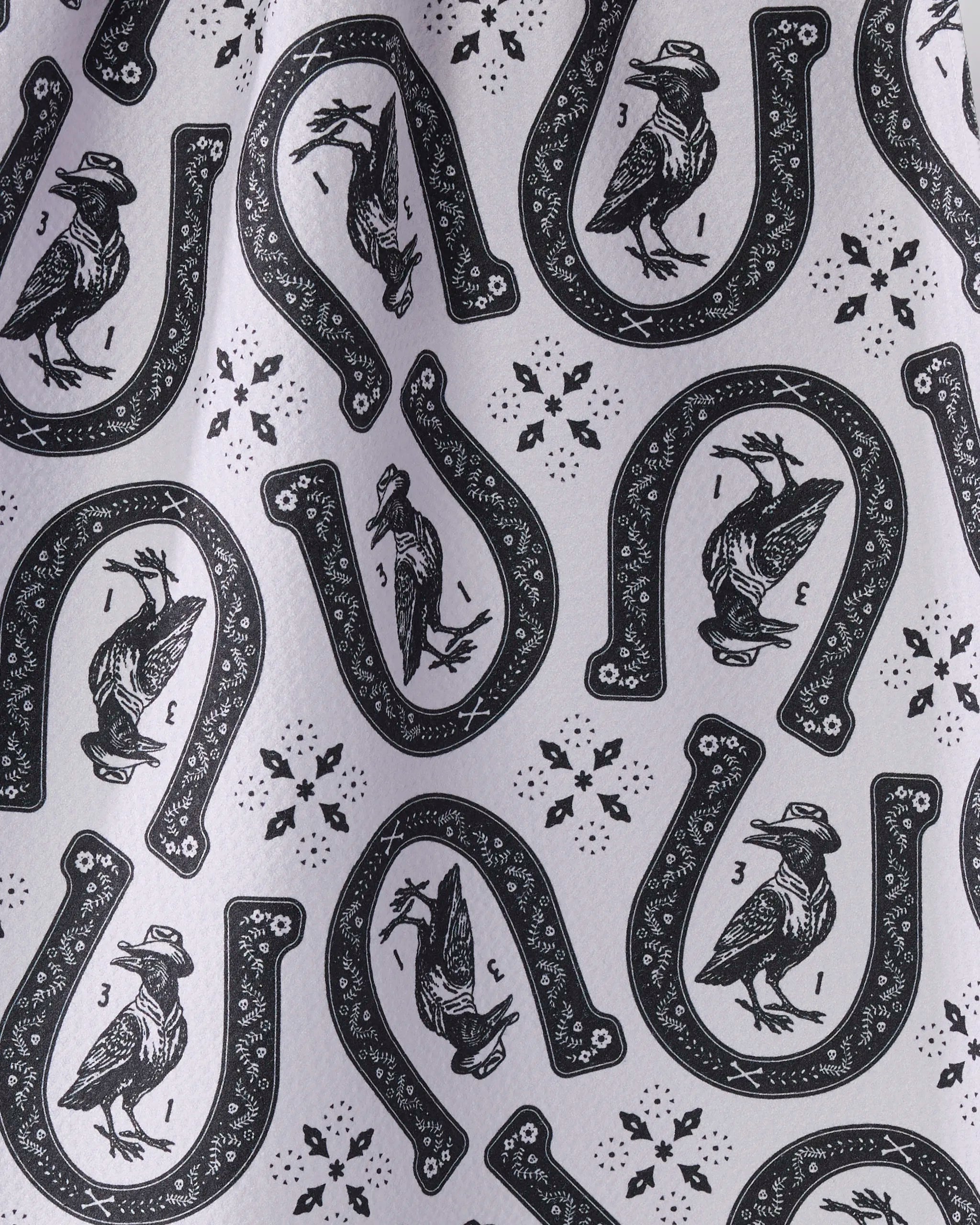 Lucky Crow by Geometry features a black and white pattern of horseshoes, hatted birds, stars, and numbers on ultra-absorbent recycled fabric. The repeating print is slightly wrinkled for textural interest.