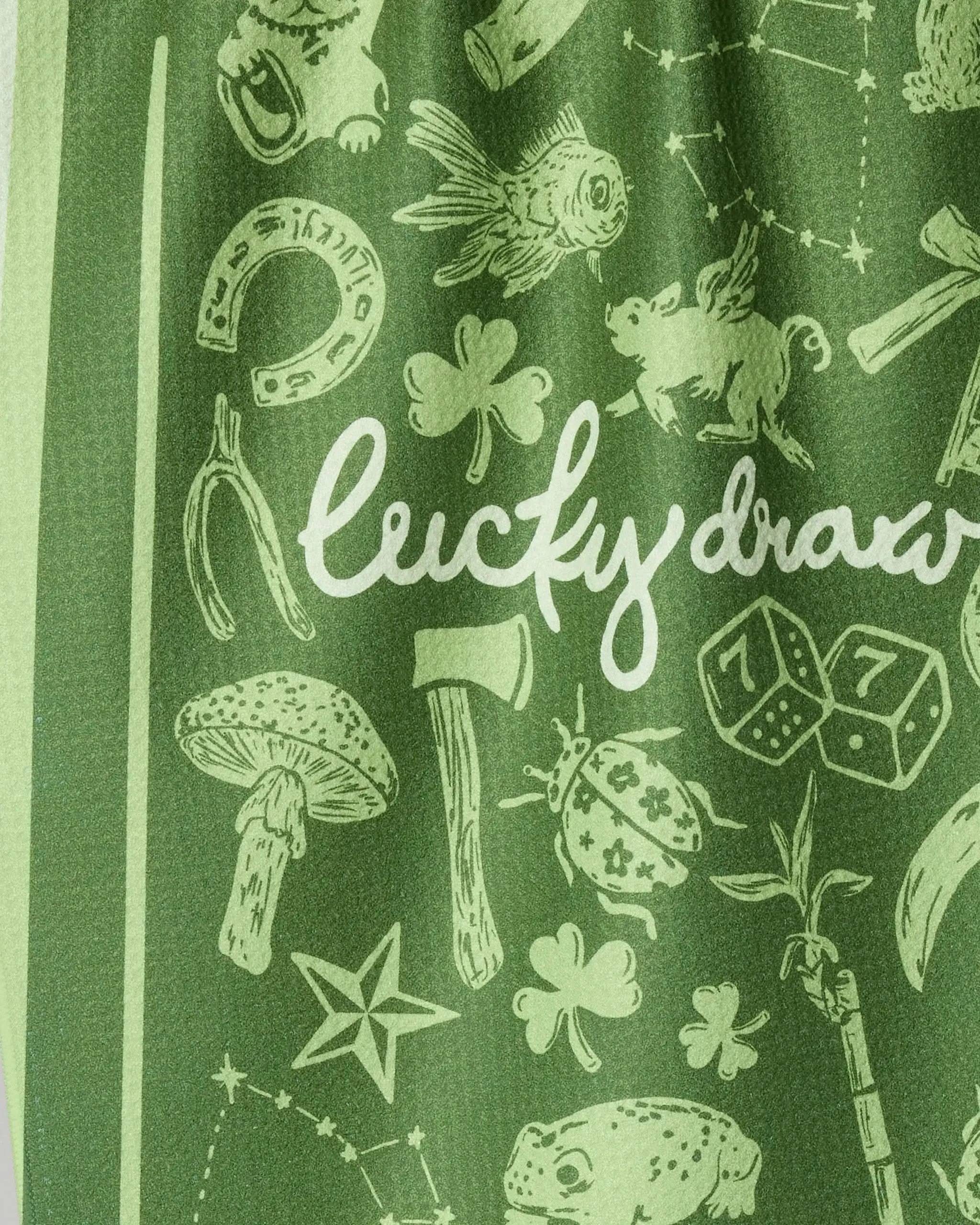 The Lucky Draw tea towel by Geometry features green eco-friendly fabric with white lucky symbols—horseshoe, four-leaf clover, pig, wishbone—and "Lucky draw" in script. Perfect as a reusable kitchen towel.