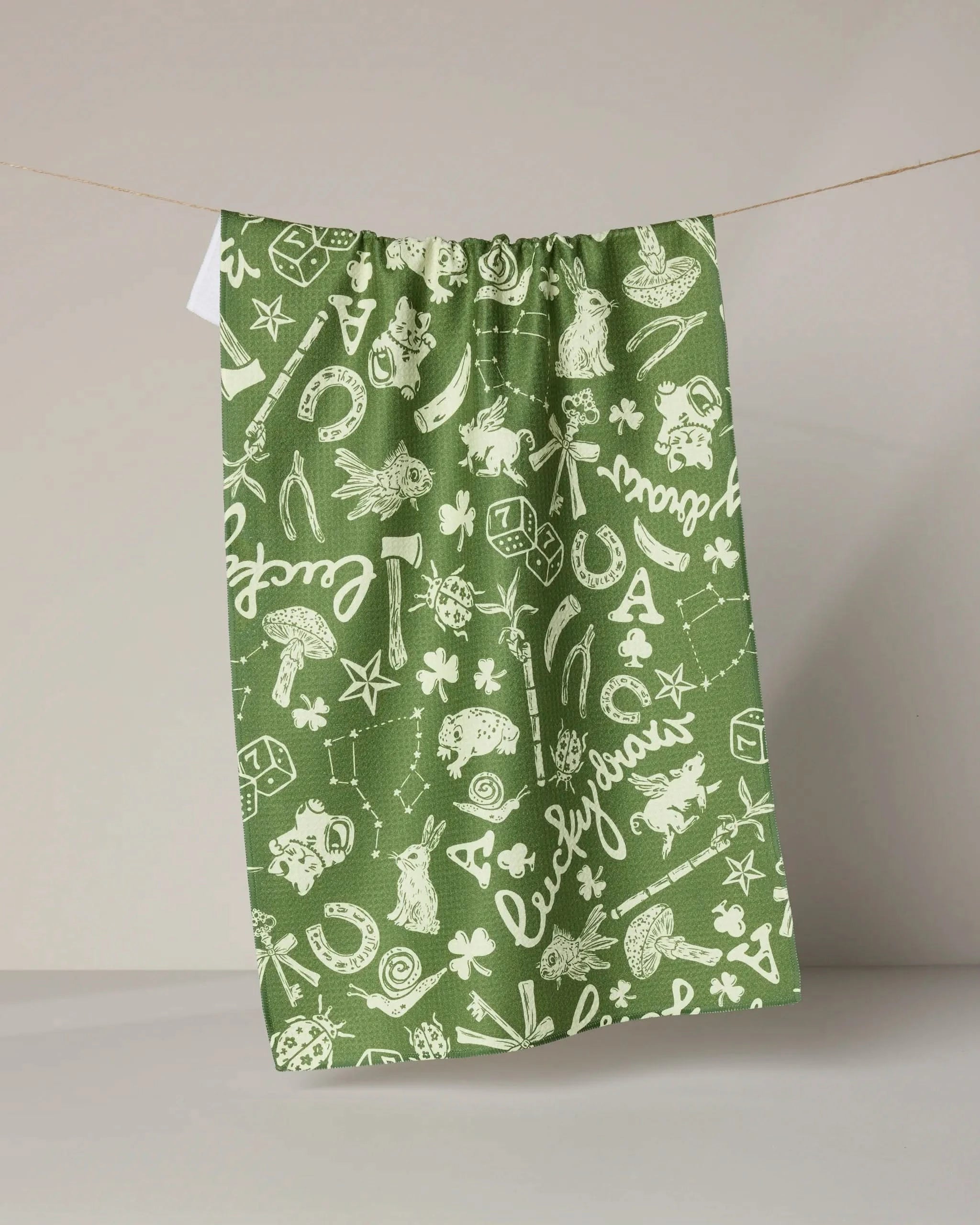Geometry’s "Lucky Draw Symbols" green tea towel is made from recycled materials and displays white whimsical illustrations—animals, stars, clovers, dice—in cursive script, shown hanging on a clothesline against a neutral background.