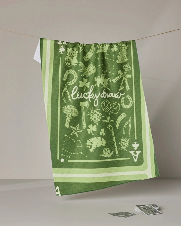 The Geometry Lucky Draw green absorbent dish towel, featuring "lucky draw" in white and decorated with luck-themed icons like horseshoes, clovers, dice, and a lucky cat, hangs over a clothesline.