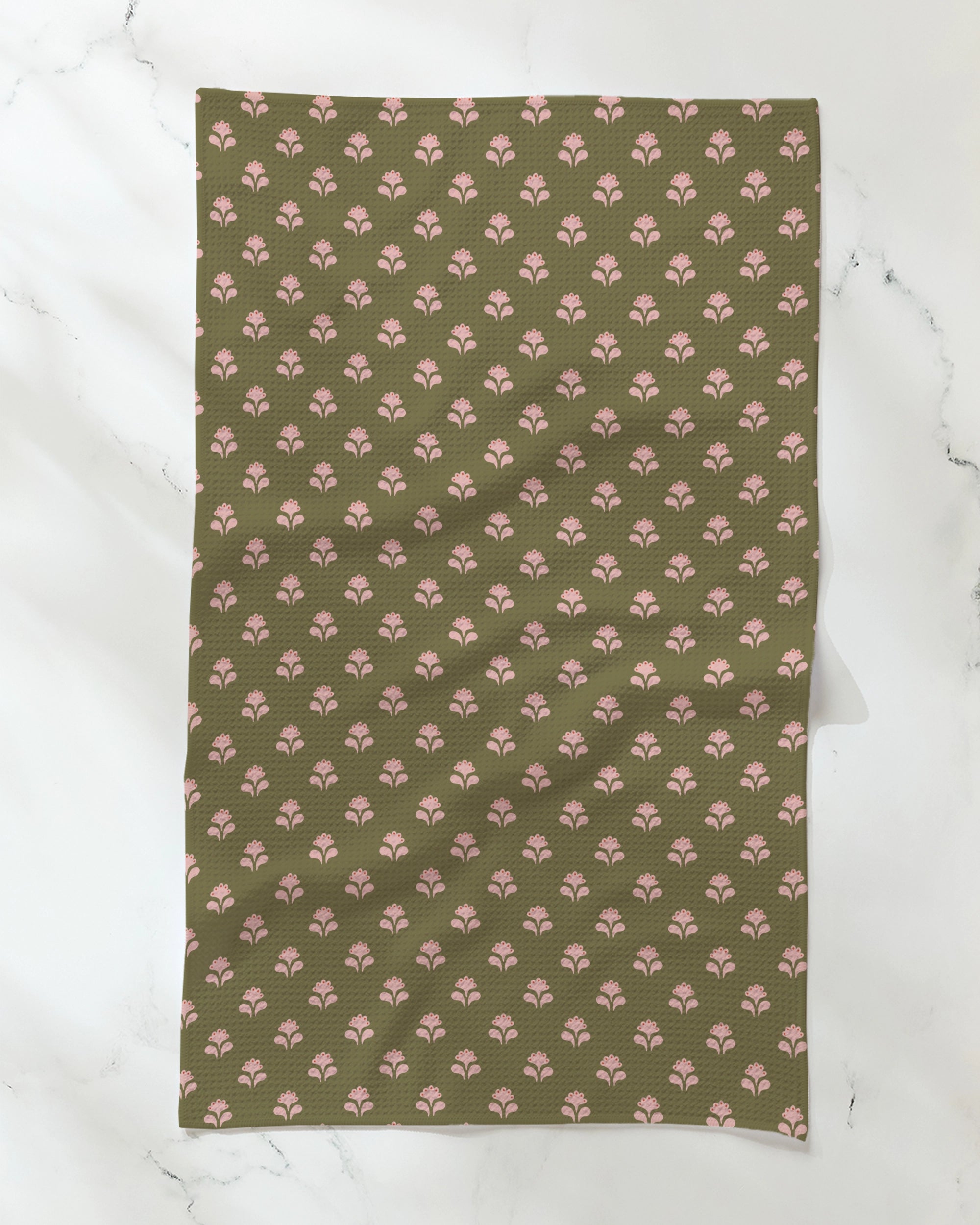Geometry’s "Lucky Flowers" tea towel showcases a rectangular design with an olive green background and a repeating pattern of small, pink fan-shaped motifs. Made from recycled materials, the towel is displayed laid flat on a marble-like white surface.
