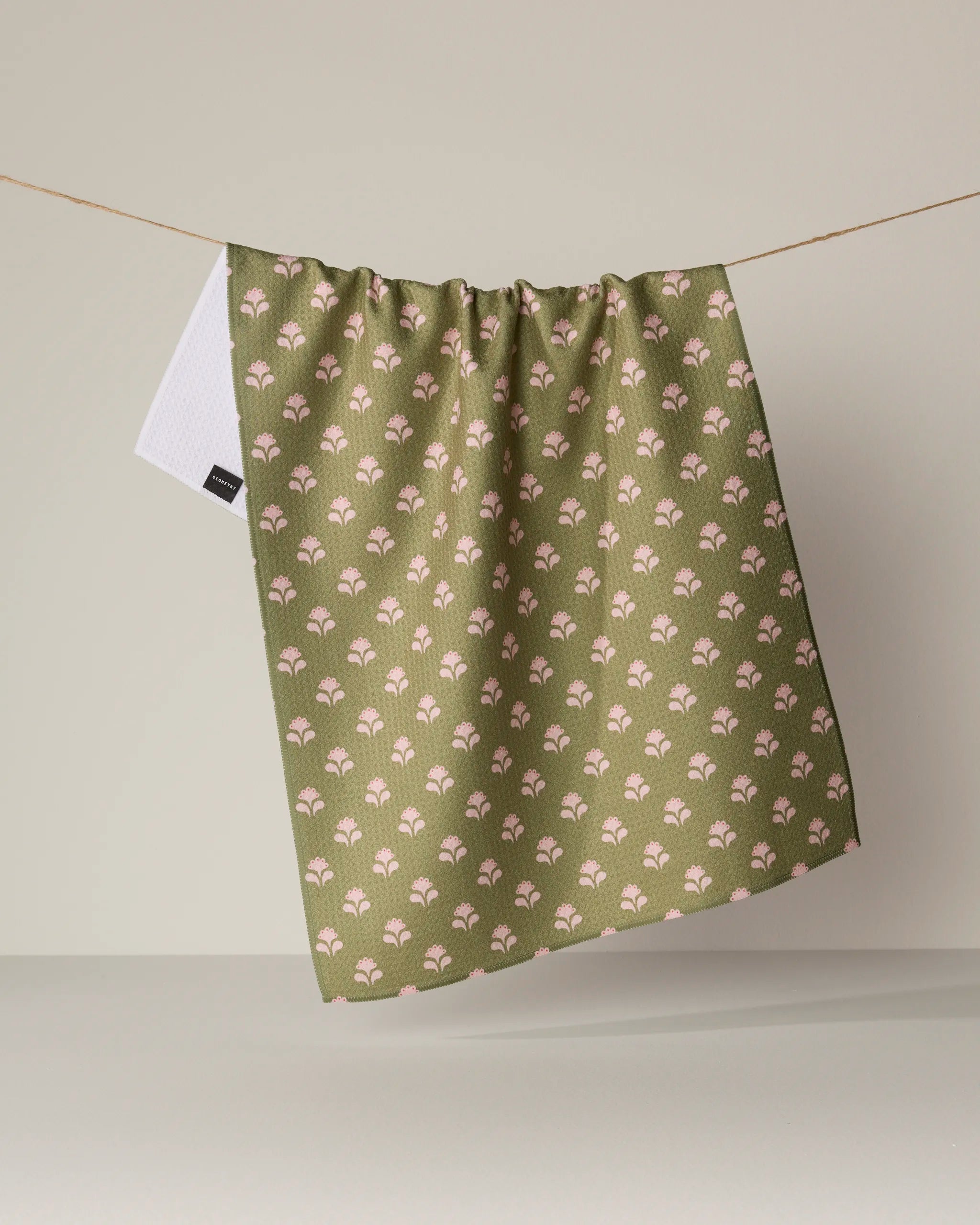 A Lucky Flowers kitchen tea towel by Geometry, featuring a small white and pink floral pattern on a green background, hangs from a string. A white tag is attached to the upper left corner of the fabric. Crafted from recycled materials, this ultra-absorbent towel is displayed against a neutral and uncluttered backdrop.