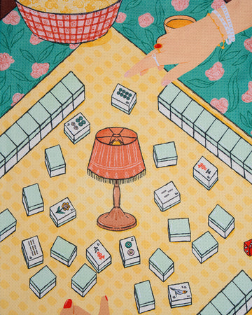 Two hands reaching over a table covered with mahjong tiles and a small lamp in the center, with a bowl of popcorn in the background atop a green floral tablecloth—accented by Geometry’s Lucky Hand kitchen towel, made from recycled materials, for an eco-friendly touch to the cozy scene.