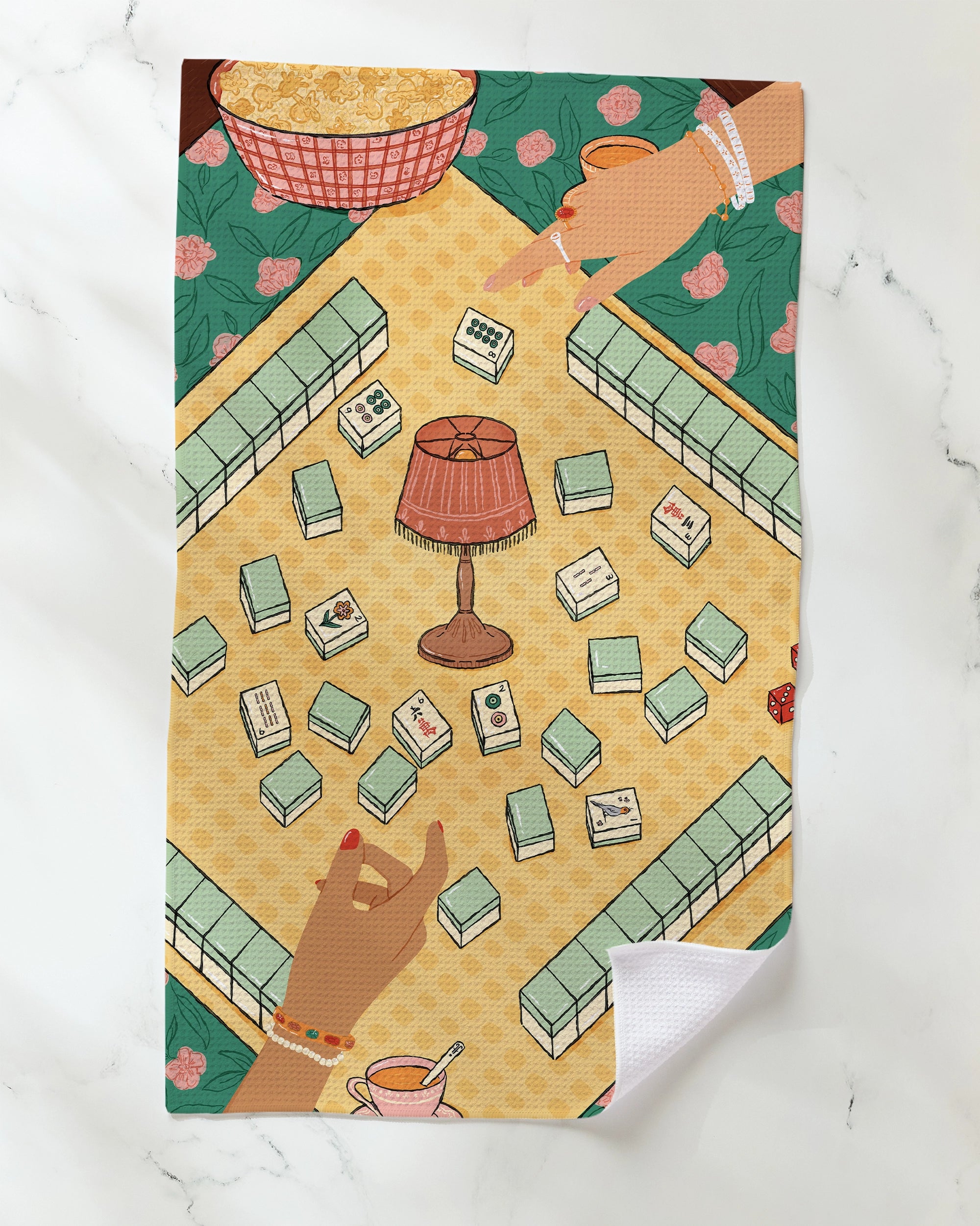 Three hands reach toward the central "Lucky Hand" lamp by Geometry, which is surrounded by scattered dominoes on a patterned tea towel. Nearby, a teacup and a bowl of popcorn add to the lively, colorful scene set on a marble surface.