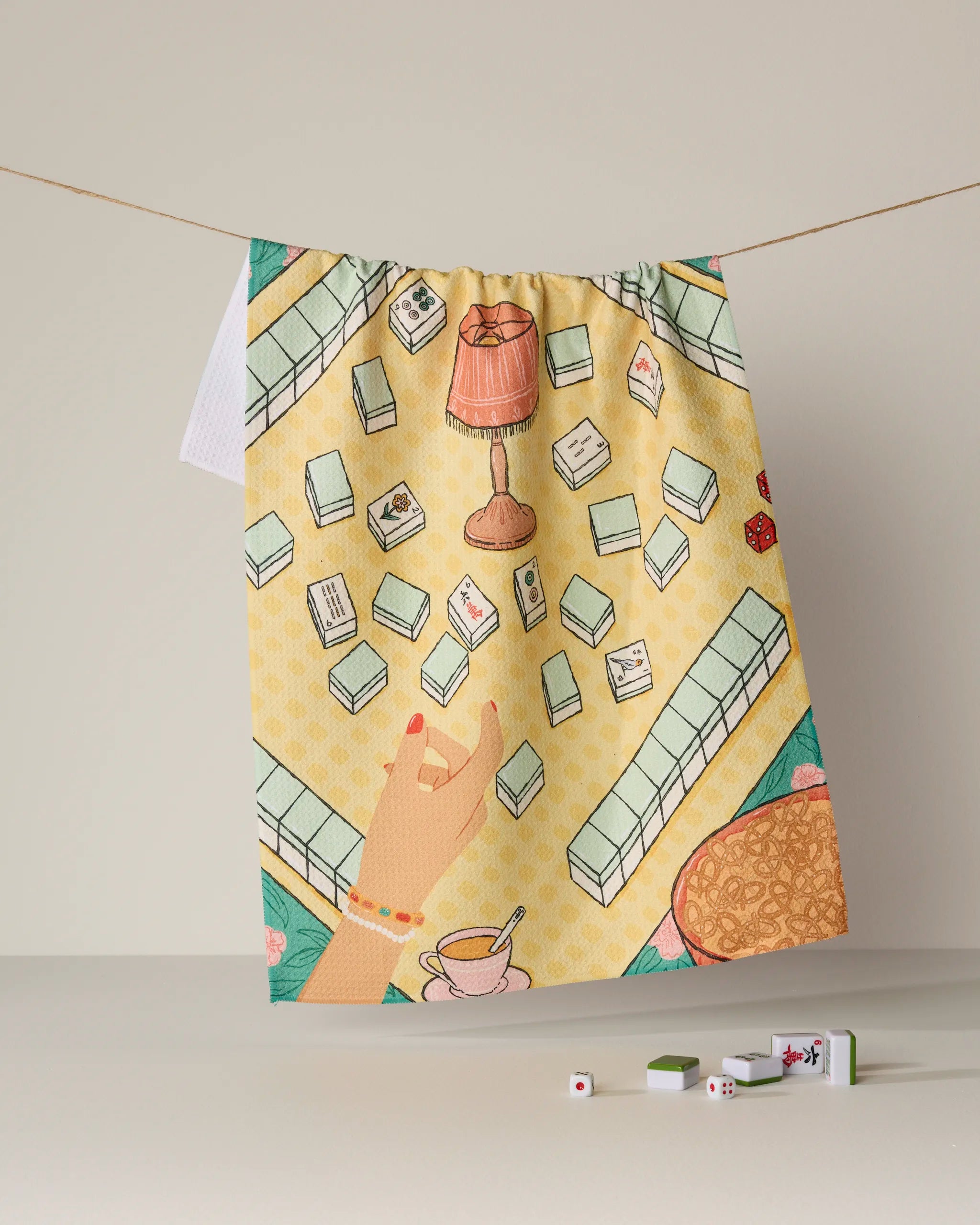 A vibrant and ultra-absorbent tea towel, "Lucky Hand" by Geometry, features a playful mahjong and tea scene illustrated against a yellow background. Mahjong tiles, a hand, a cup of tea, and a lamp adorn this eco-friendly towel crafted from recycled materials. Both practical and charming, it hangs on a clothesline with real mahjong tiles and dice placed below for added detail.