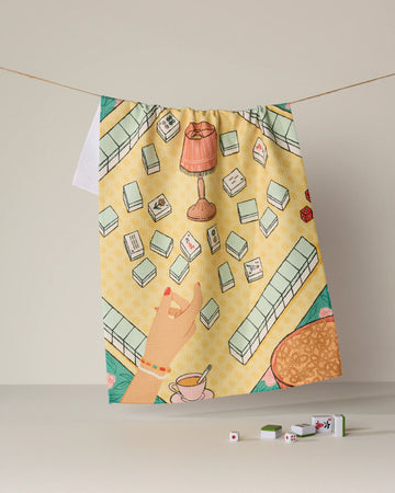 A vibrant and ultra-absorbent tea towel, "Lucky Hand" by Geometry, features a playful mahjong and tea scene illustrated against a yellow background. Mahjong tiles, a hand, a cup of tea, and a lamp adorn this eco-friendly towel crafted from recycled materials. Both practical and charming, it hangs on a clothesline with real mahjong tiles and dice placed below for added detail.