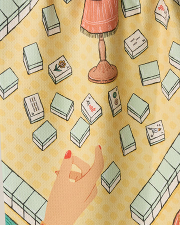 A hand reaches toward a Mahjong tile on a table with scattered tiles, a lamp, and the Geometry Lucky Hand patterned yellow tea towel made from ultra-absorbent recycled materials.