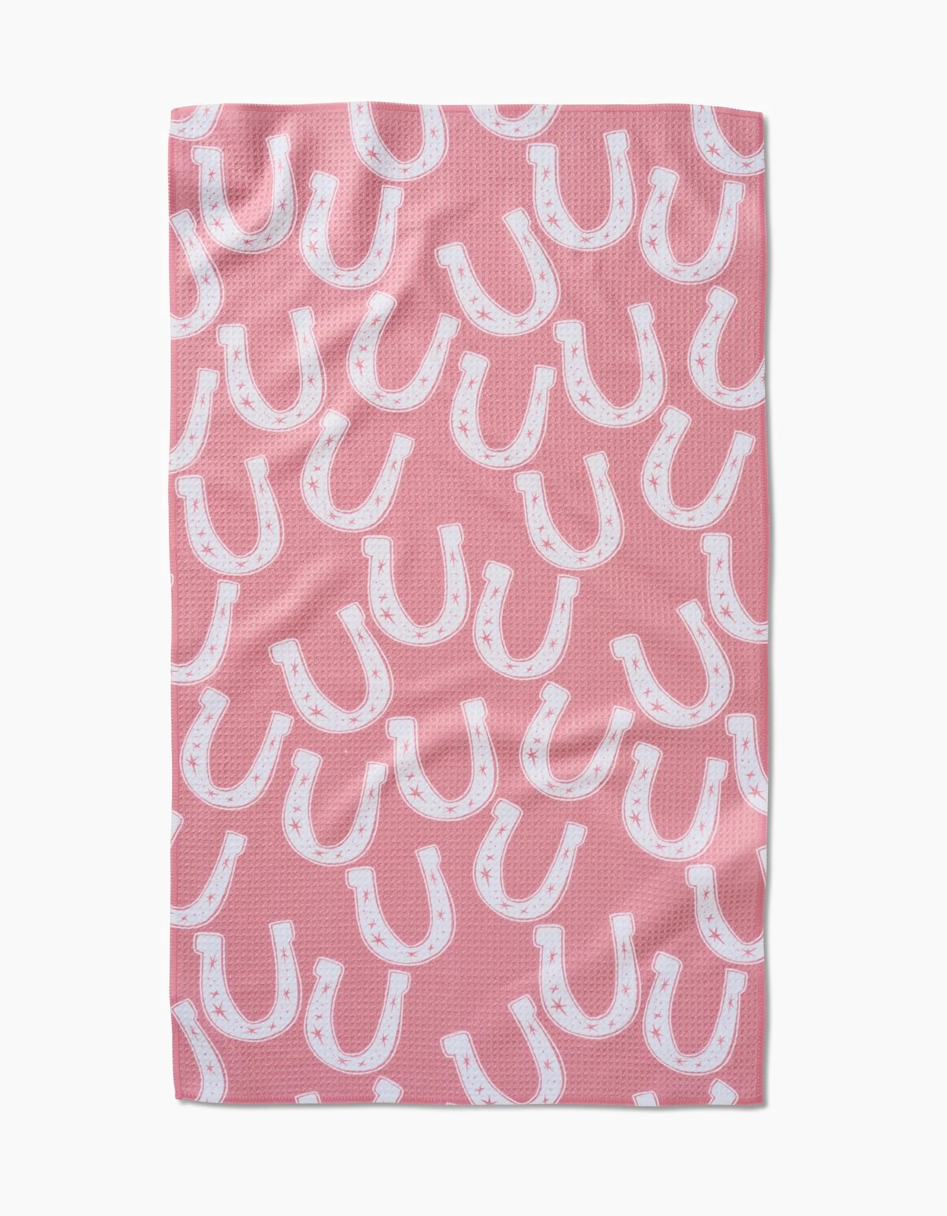 The Lucky Horseshoe tea towel by Aubrey Fairchild features a pink Geoweave fabric with a repeating pattern of white horseshoes outlined in black and accented with small pink stars.