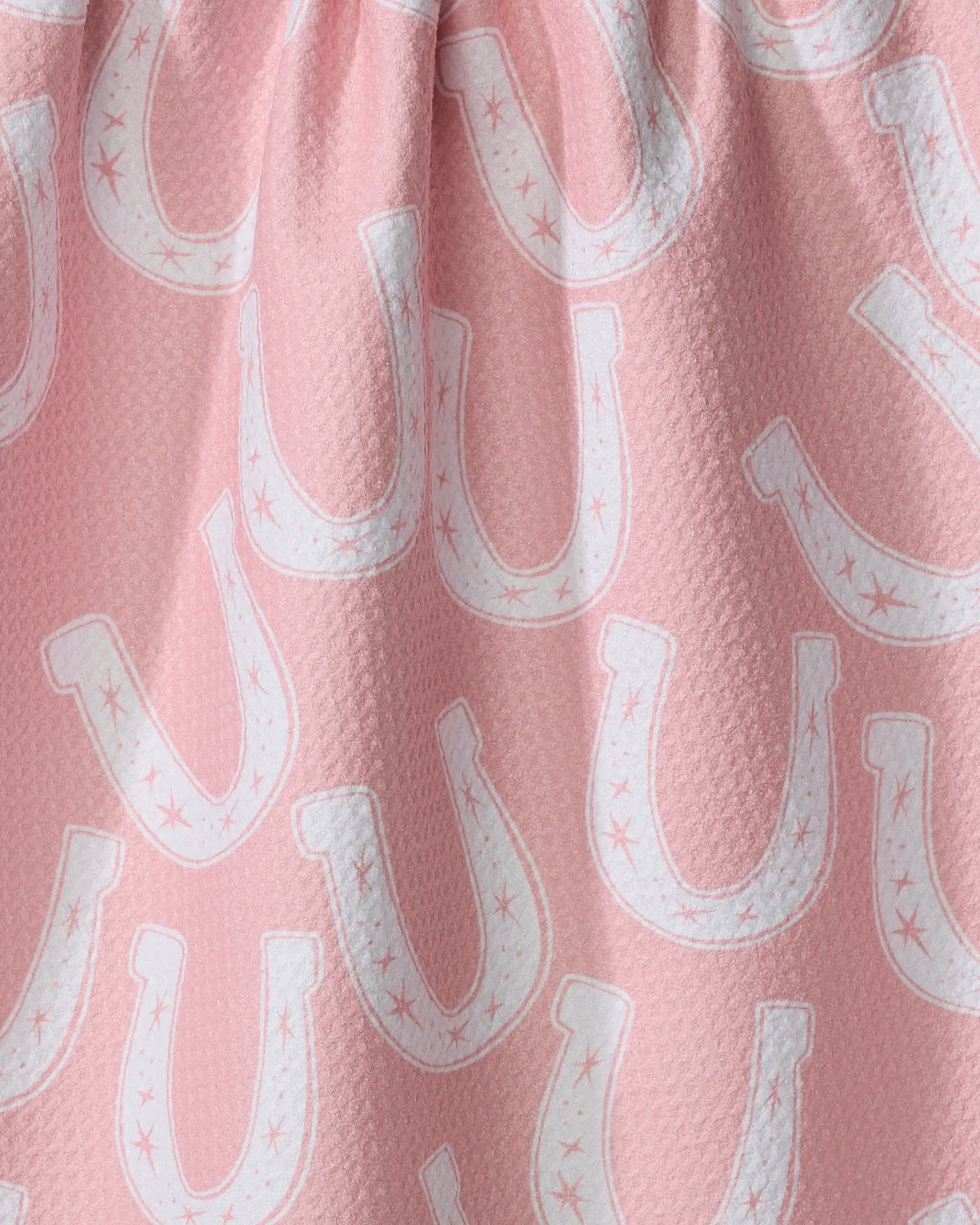 The Lucky Horseshoe tea towel by Aubrey Fairchild features a pink background with white horseshoes, some accented with stars. Its subtly textured fabric and gentle folds make it both appealing and practical for everyday use.