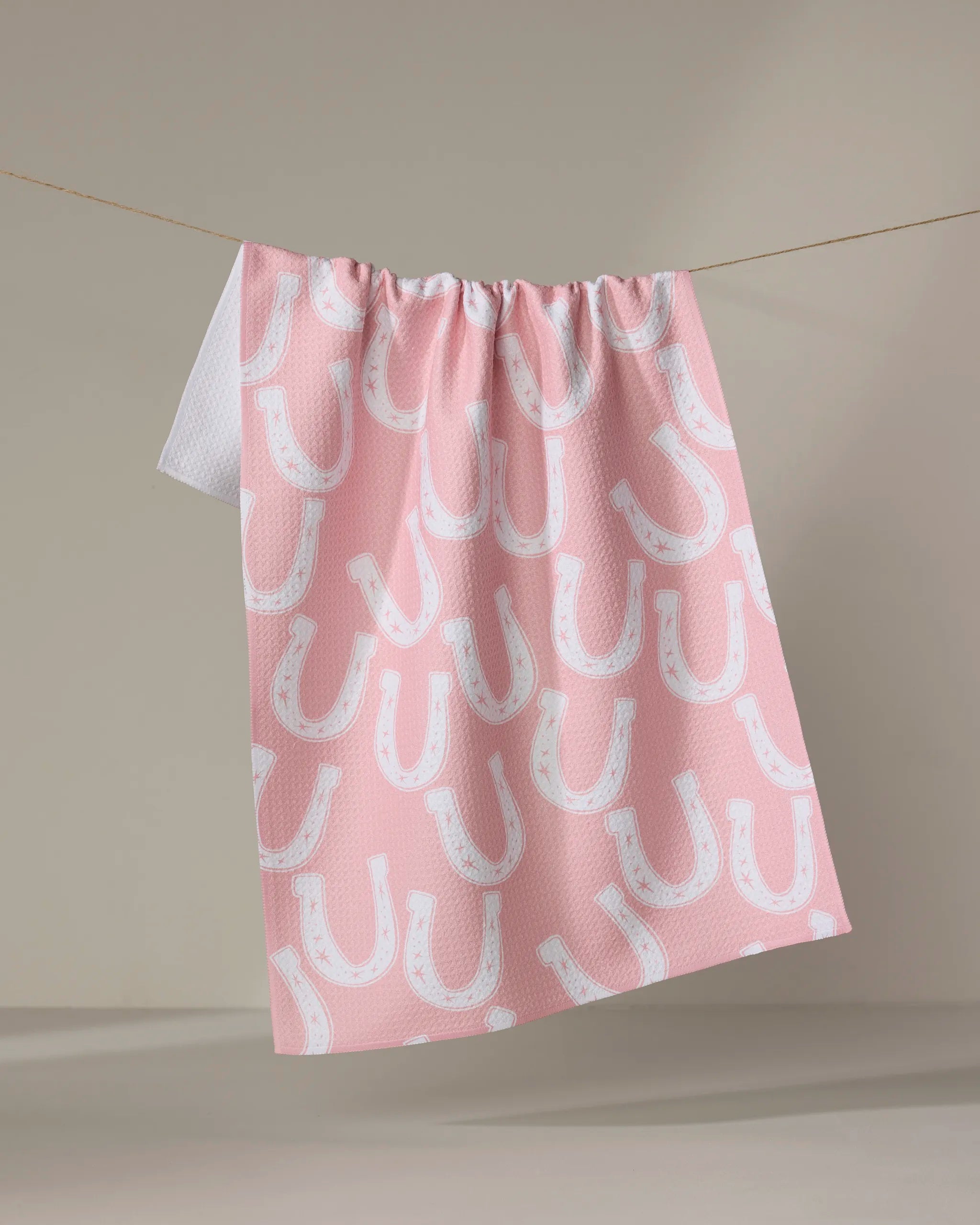 The Lucky Horseshoe towel by Aubrey Fairchild, quick-drying and pink with a white horseshoe pattern, hangs on a clothesline against a neutral background.