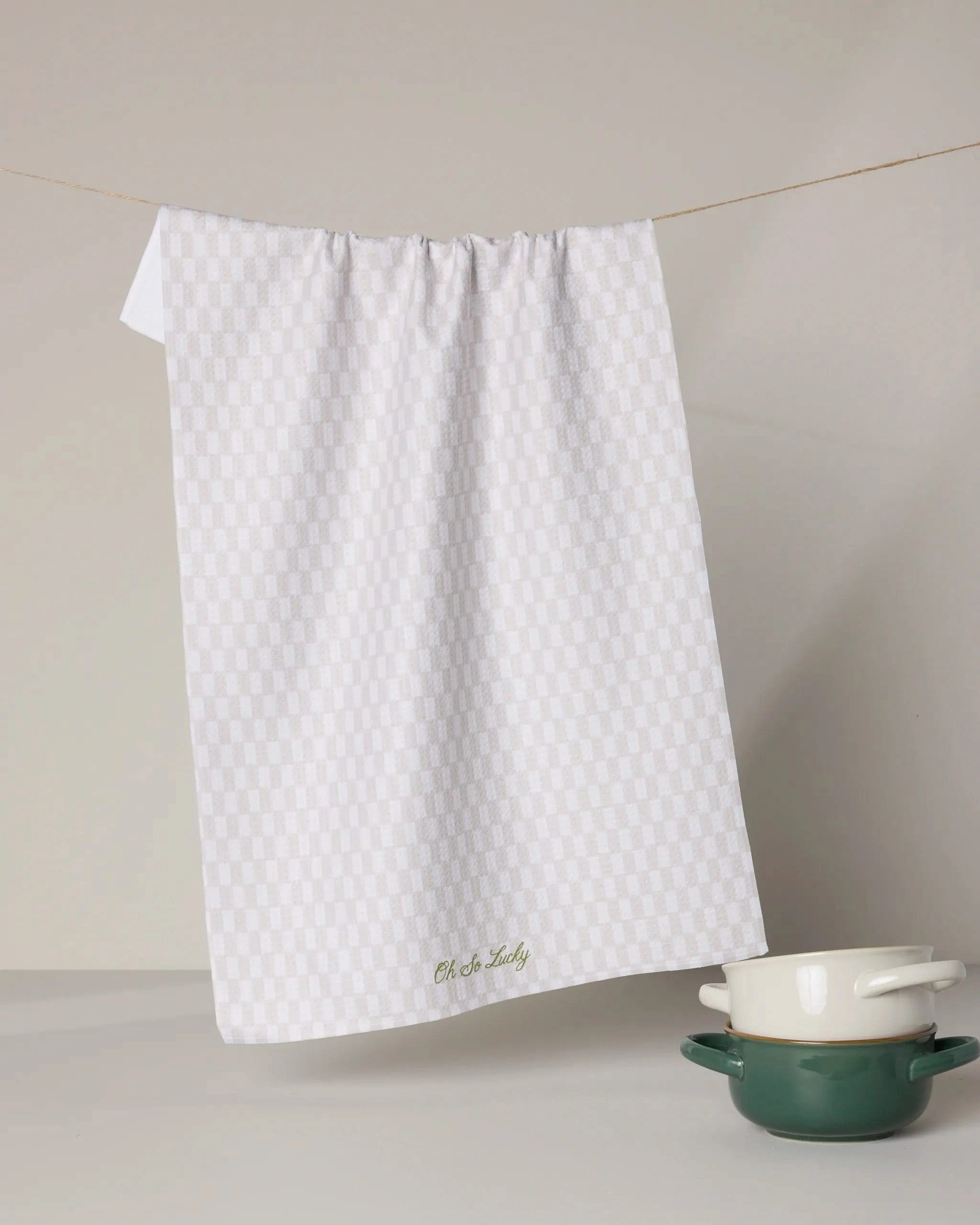 A white, ultra-absorbent Geometry Lucky Lilah tea towel hangs on a string against a neutral background, with a green pot featuring a white lid and a matching small bowl arranged below on a flat surface.