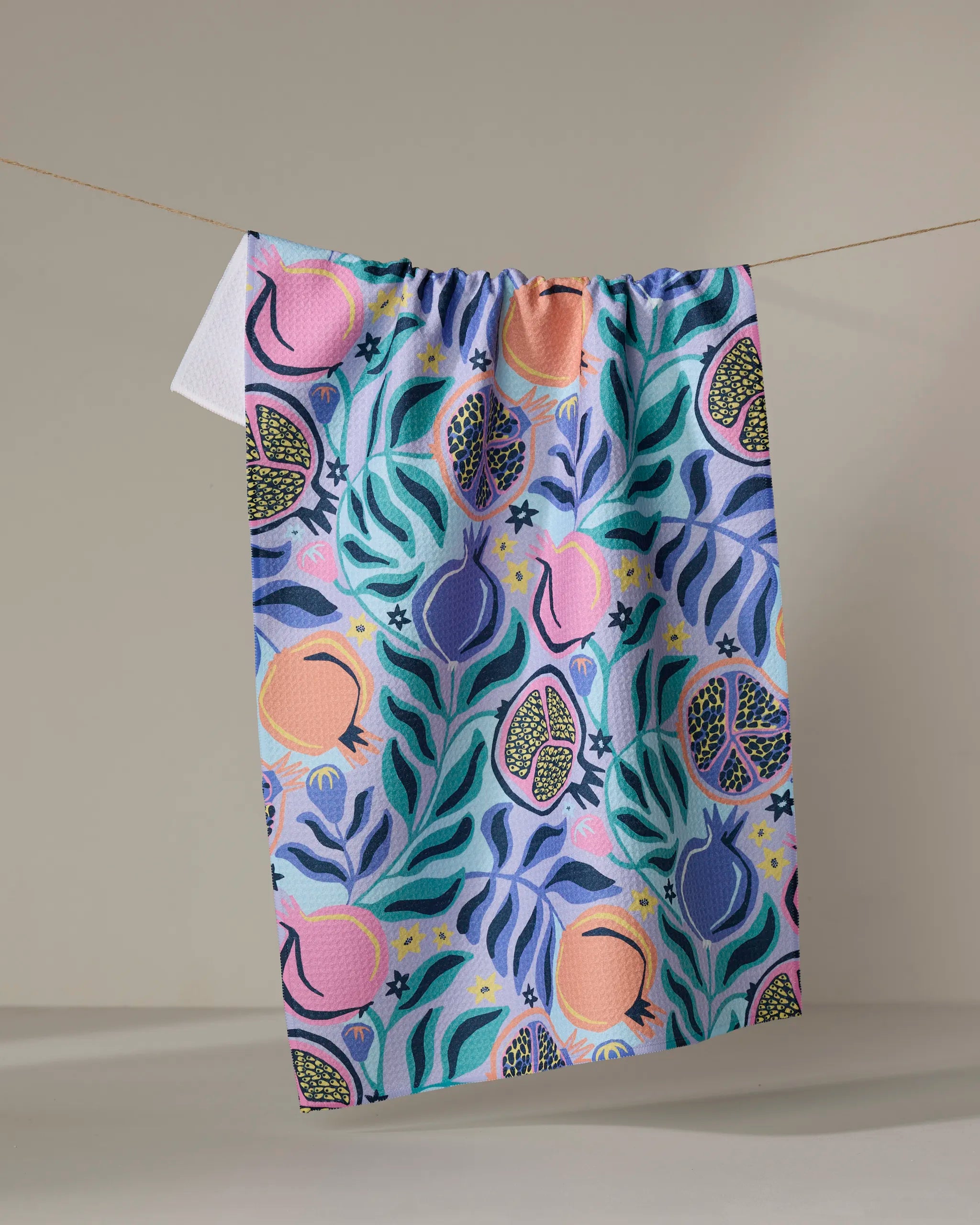 The Lucky Pomegranate kitchen towel by Masha Volnova, made from recycled materials, features a vibrant abstract fruit and leaf design in pink, blue, green, and yellow, hanging against a plain beige background.