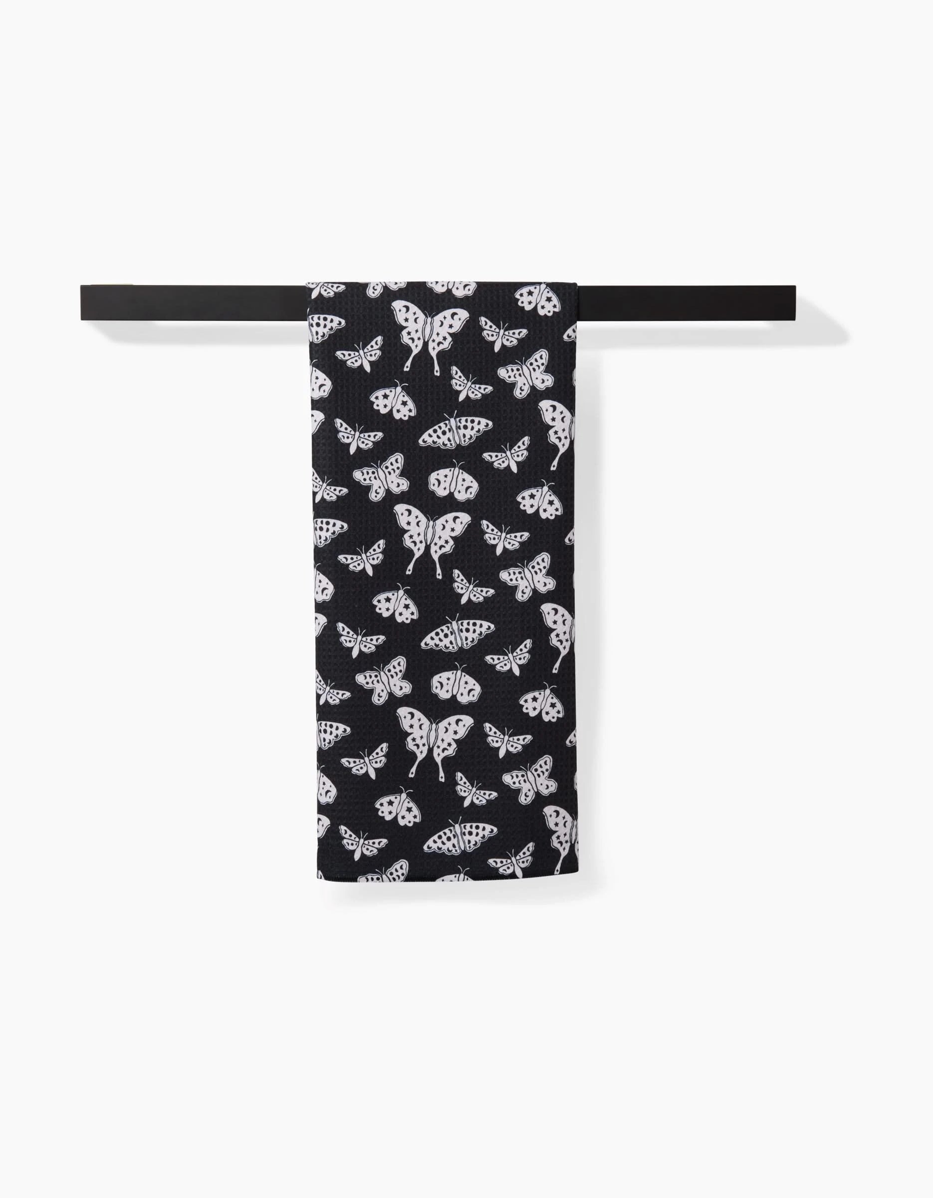 The Kayla Ann Lunar Moths Black towel, featuring a white moth pattern, is neatly folded over a minimalist black bar against a white background.