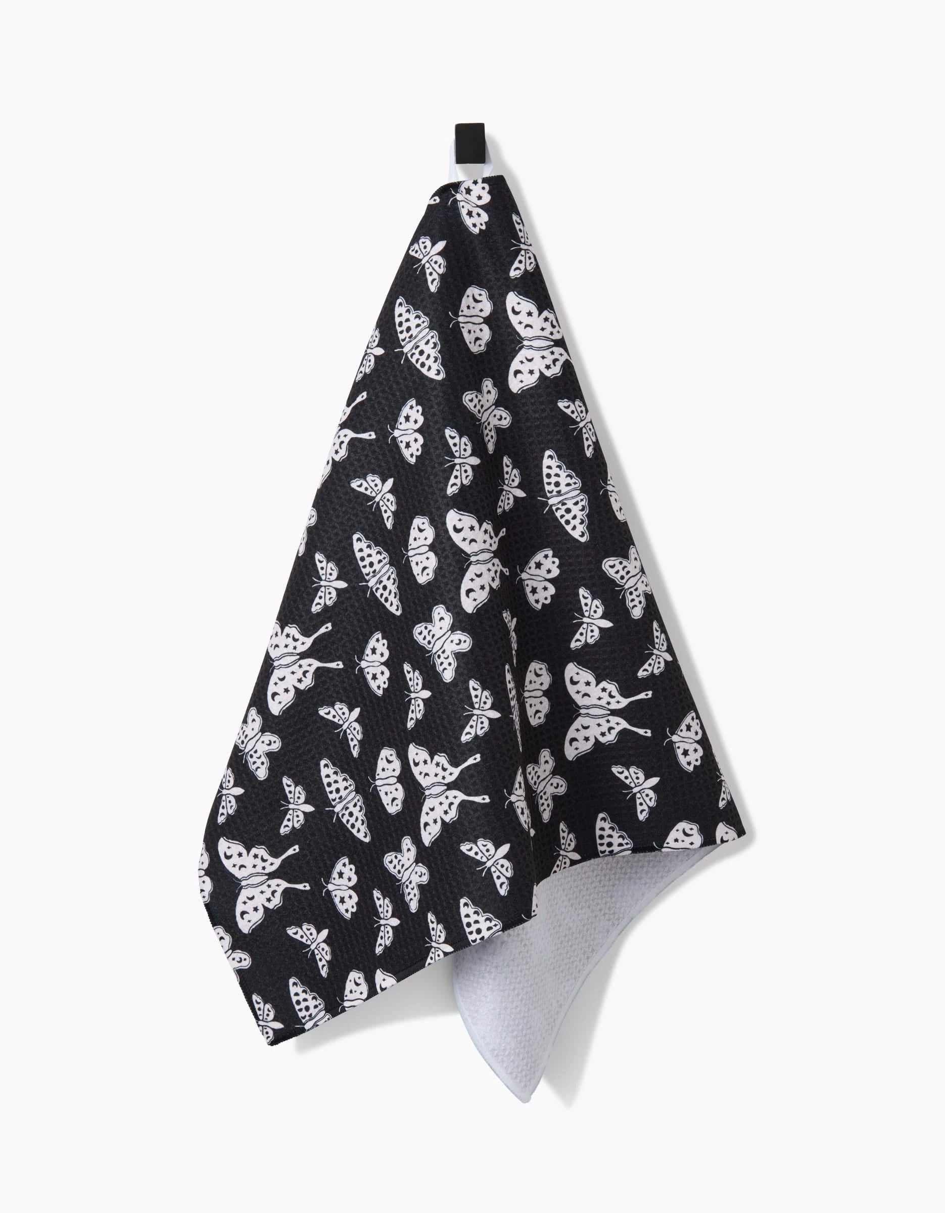 The Kayla Ann Lunar Moths Black recycled kitchen towel displays white moth and butterfly patterns on black fabric, with a top black hanging loop, shown against a white background.